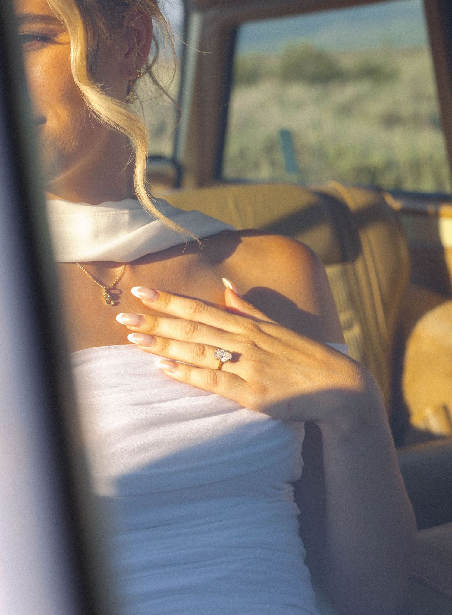 Bride in white strapless gown displaying diamond engagement ring while seated in vintage car interior.