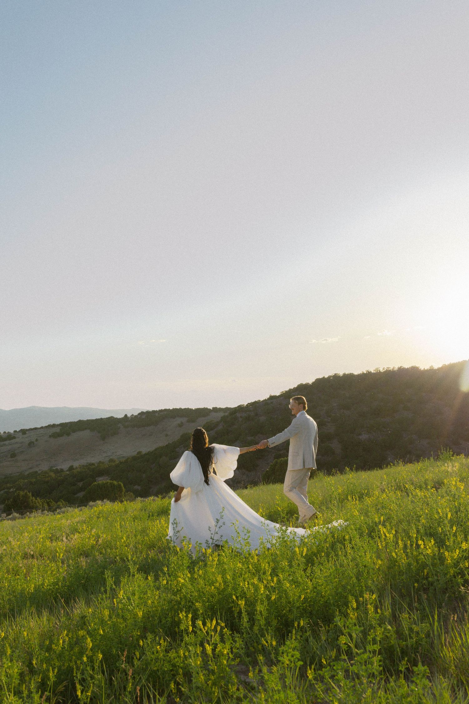 Bride and groom holding hands walking through green meadow at sunset with rolling hills in background.