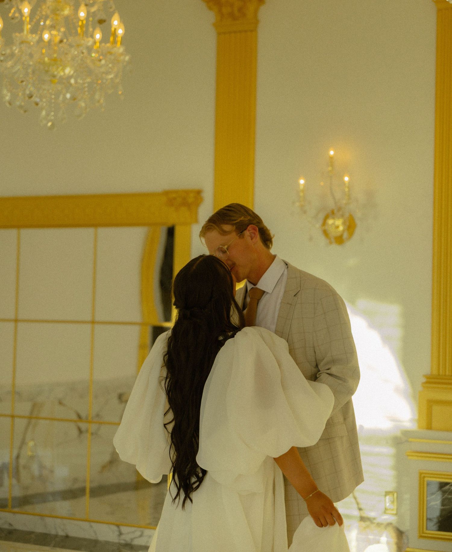 Couple sharing a romantic kiss in an elegant venue with crystal chandelier and gold accents during their wedding ceremony.