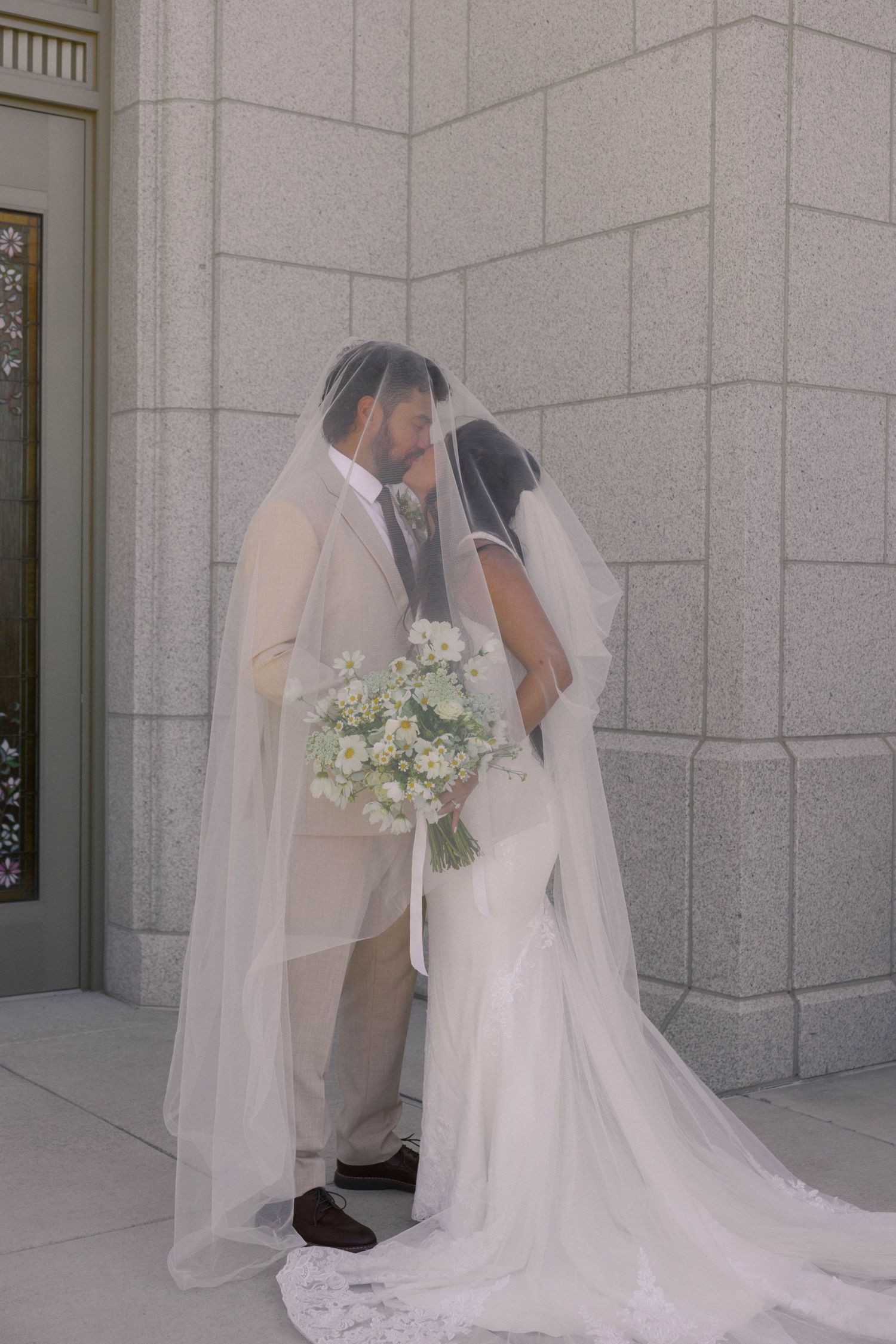 Bride and groom kissing under a flowing white veil, holding a white bouquet, standing against a marble wall.