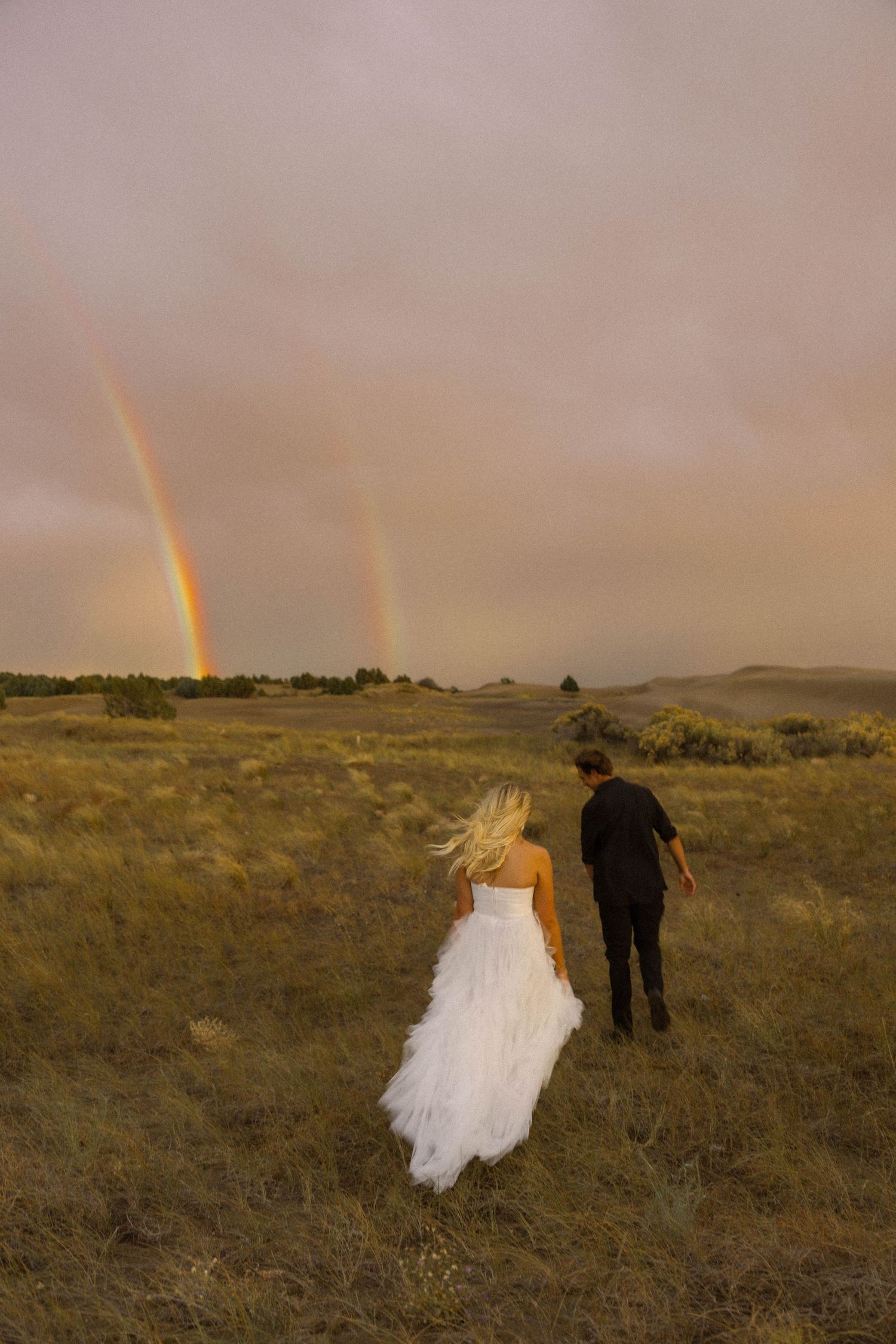 Bride and groom walking through golden fields toward a stunning double rainbow under dramatic stormy skies.