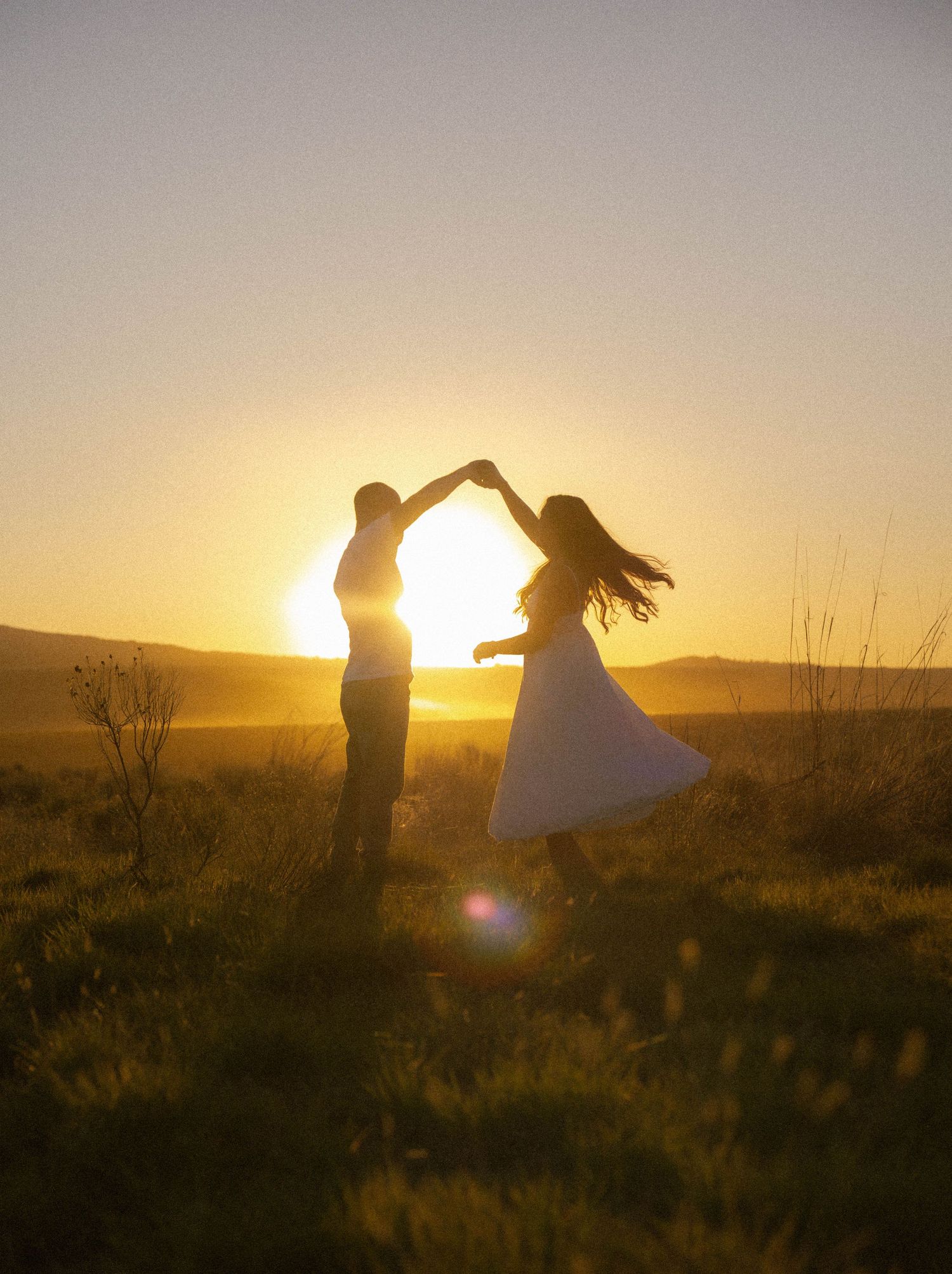 Couple dancing in silhouette against golden sunset in open field, woman in white dress twirling gracefully.