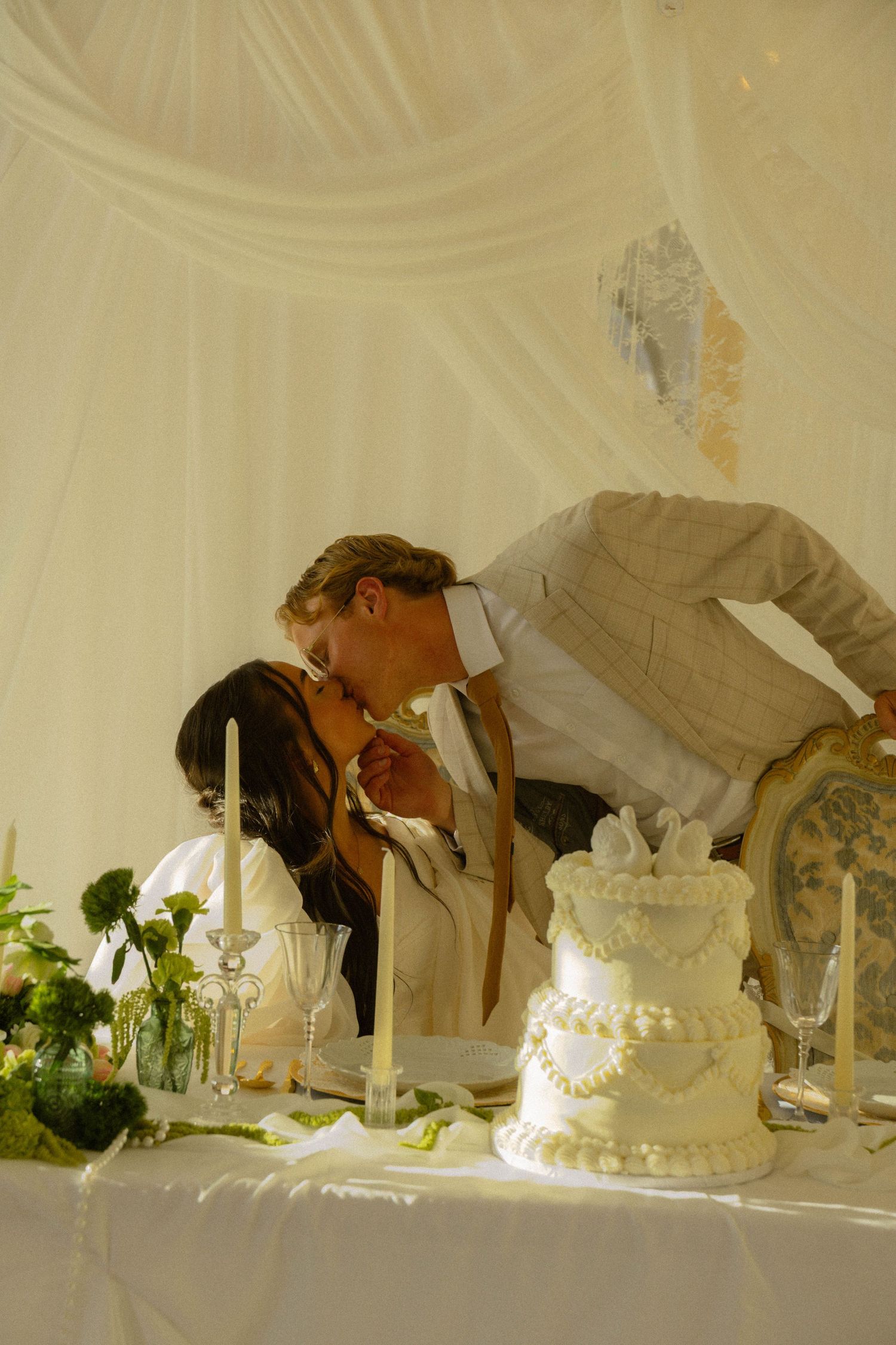 Bride and groom sharing a kiss at wedding reception table with white tiered cake and candles under draped tent.