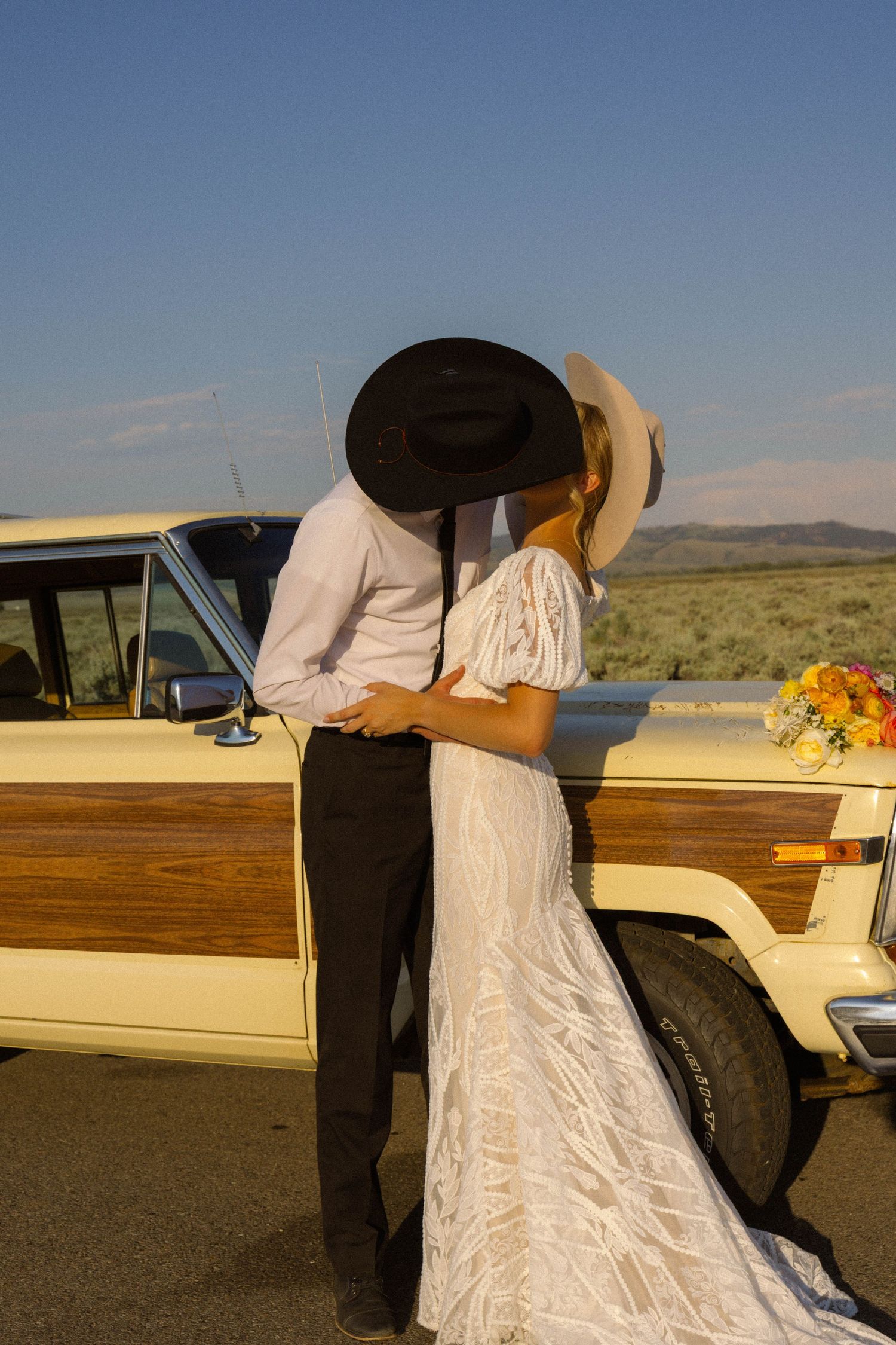 Bride and groom in cowboy hats kissing beside a vintage woody wagon in a desert landscape.