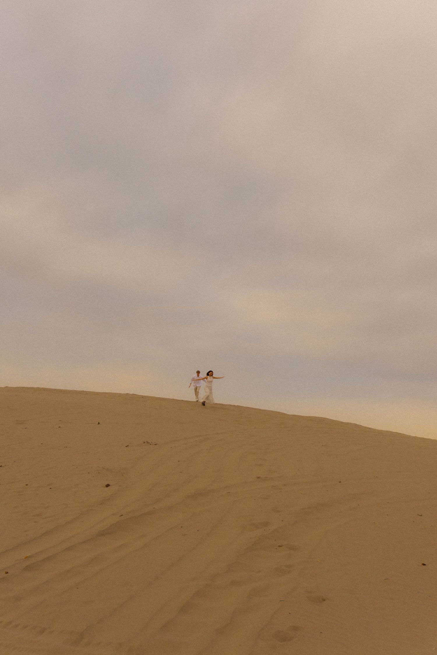 Two people in white clothing stand atop a vast sandy desert dune under an overcast sky.