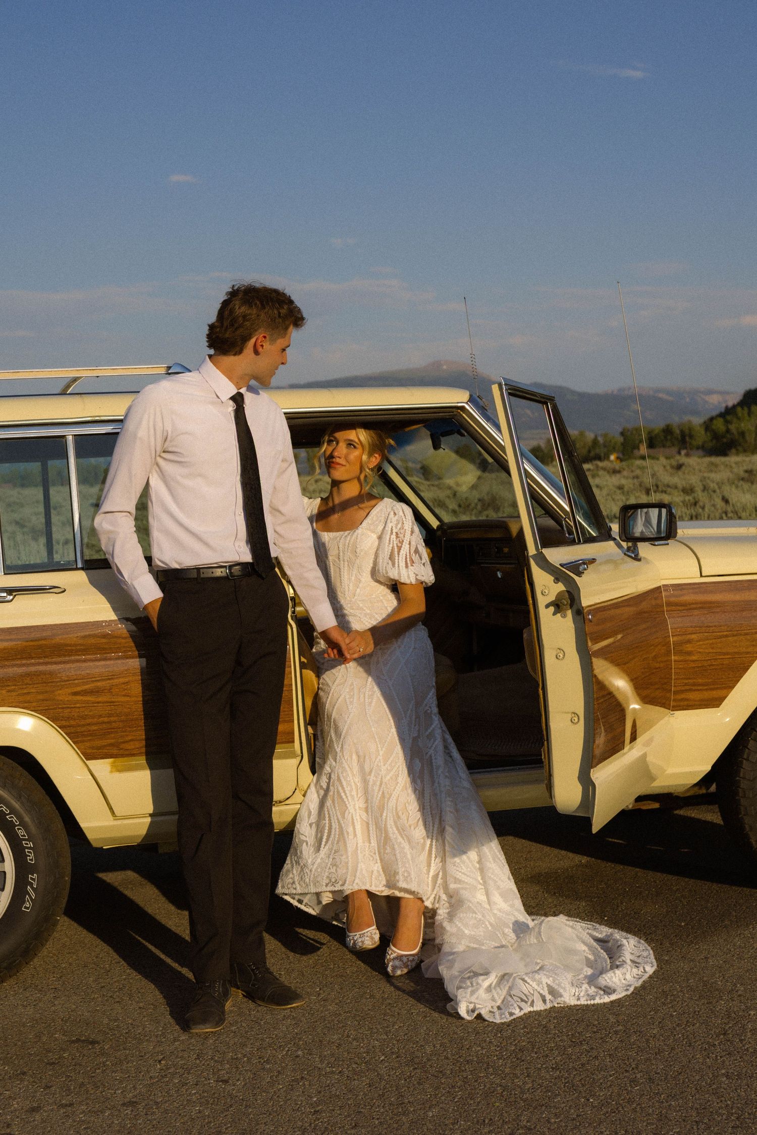 Bride in white lace dress and groom in white shirt and tie pose by a vintage woody wagon outdoors.