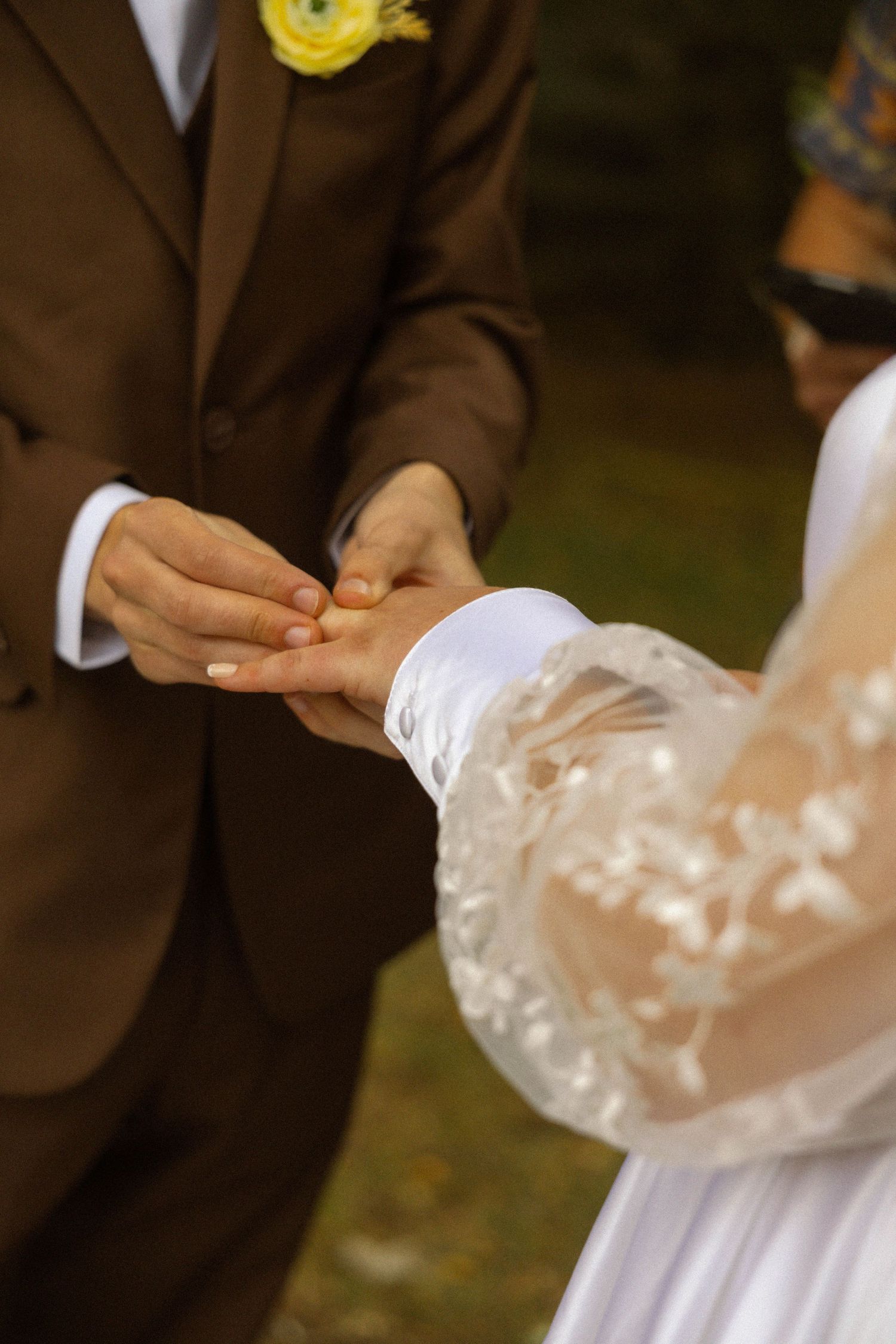 Groom in brown suit placing wedding ring on bride's finger during outdoor ceremony.
