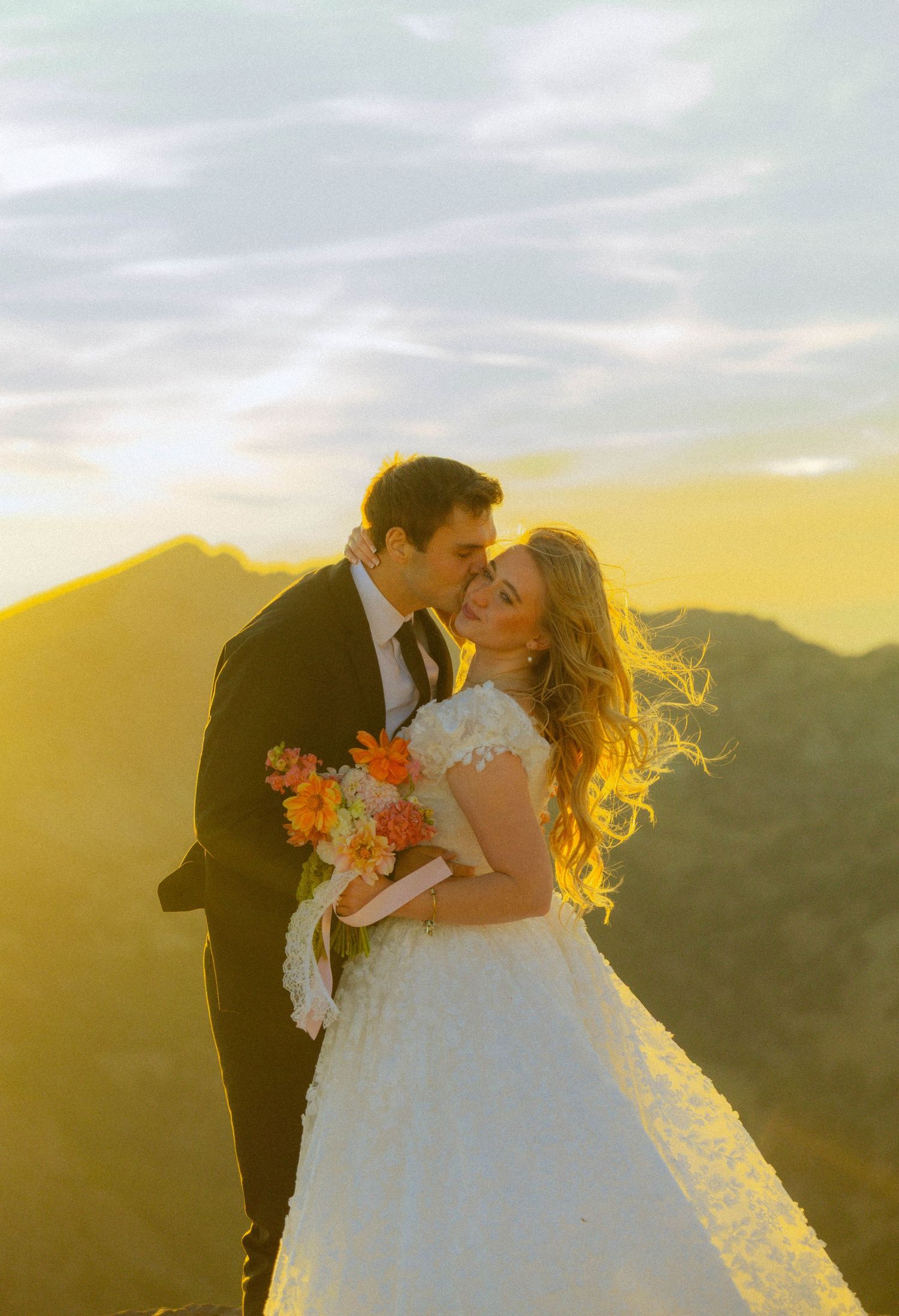 Bride and groom share a romantic kiss at golden sunset on mountain peak, bride holding vibrant orange bouquet in white lace gown.