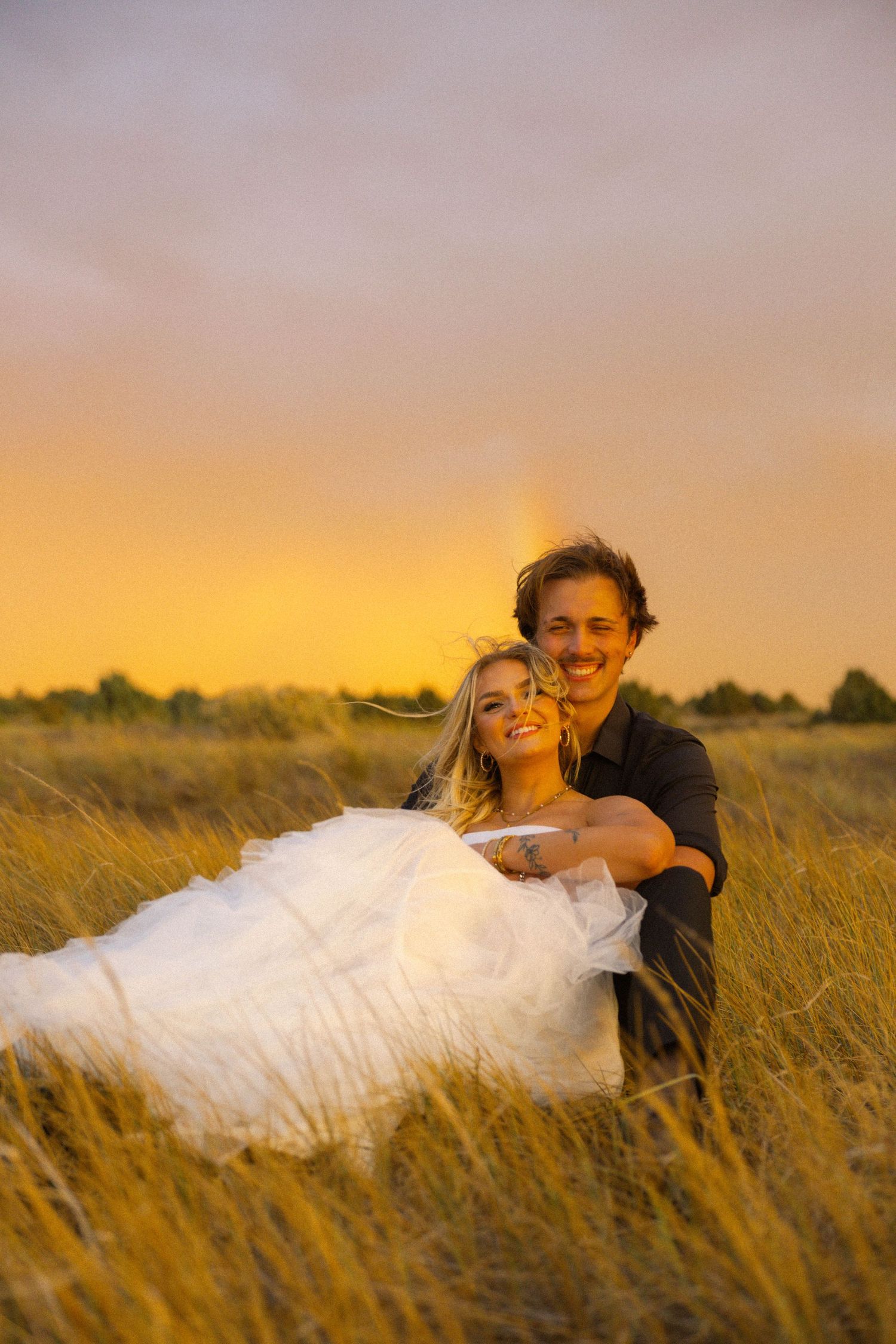 A groom holds his smiling bride in a golden wheat field at sunset during their romantic outdoor wedding photo session.