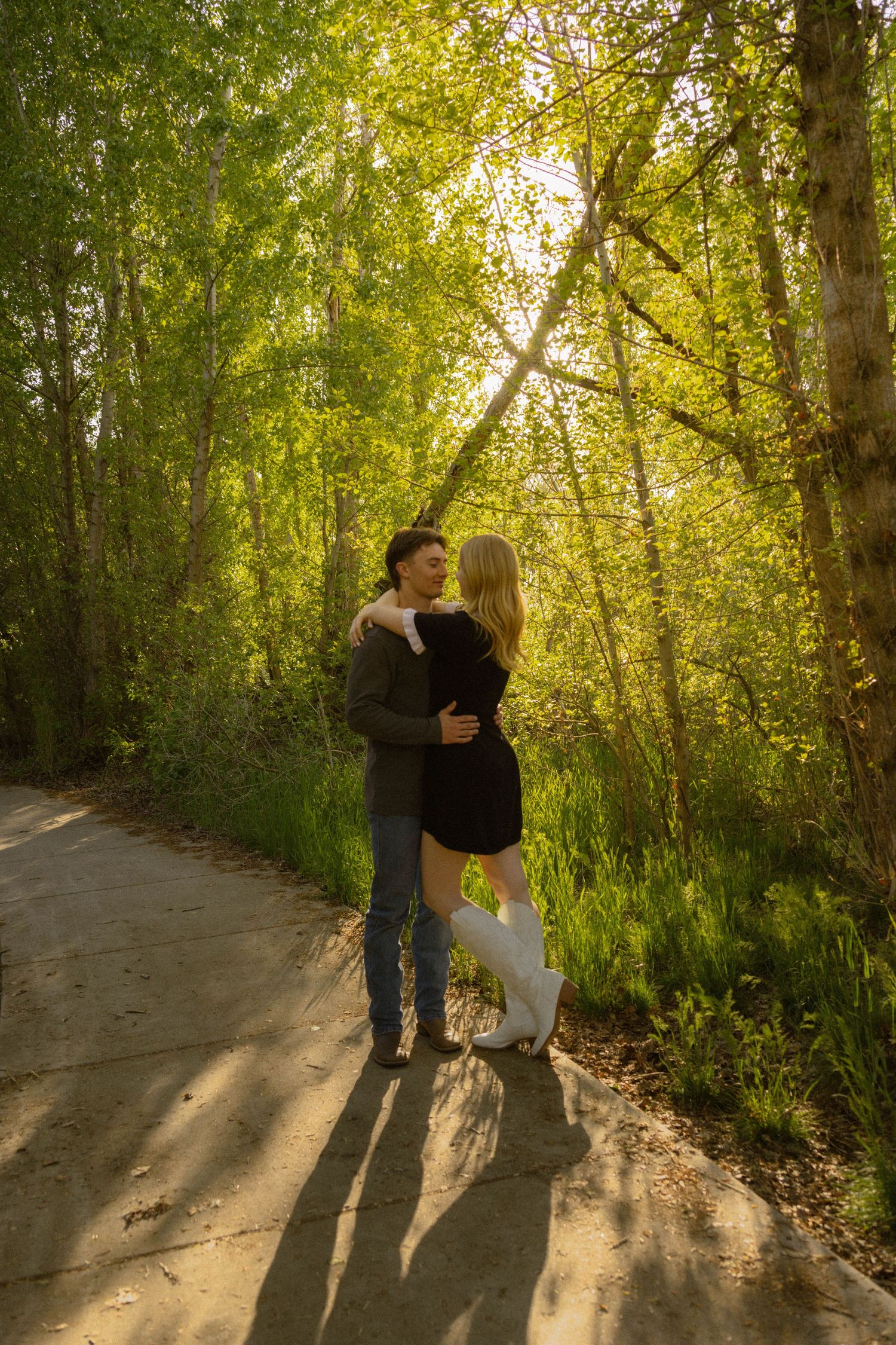 Couple embracing on a sunlit forest path surrounded by lush green trees during golden hour.