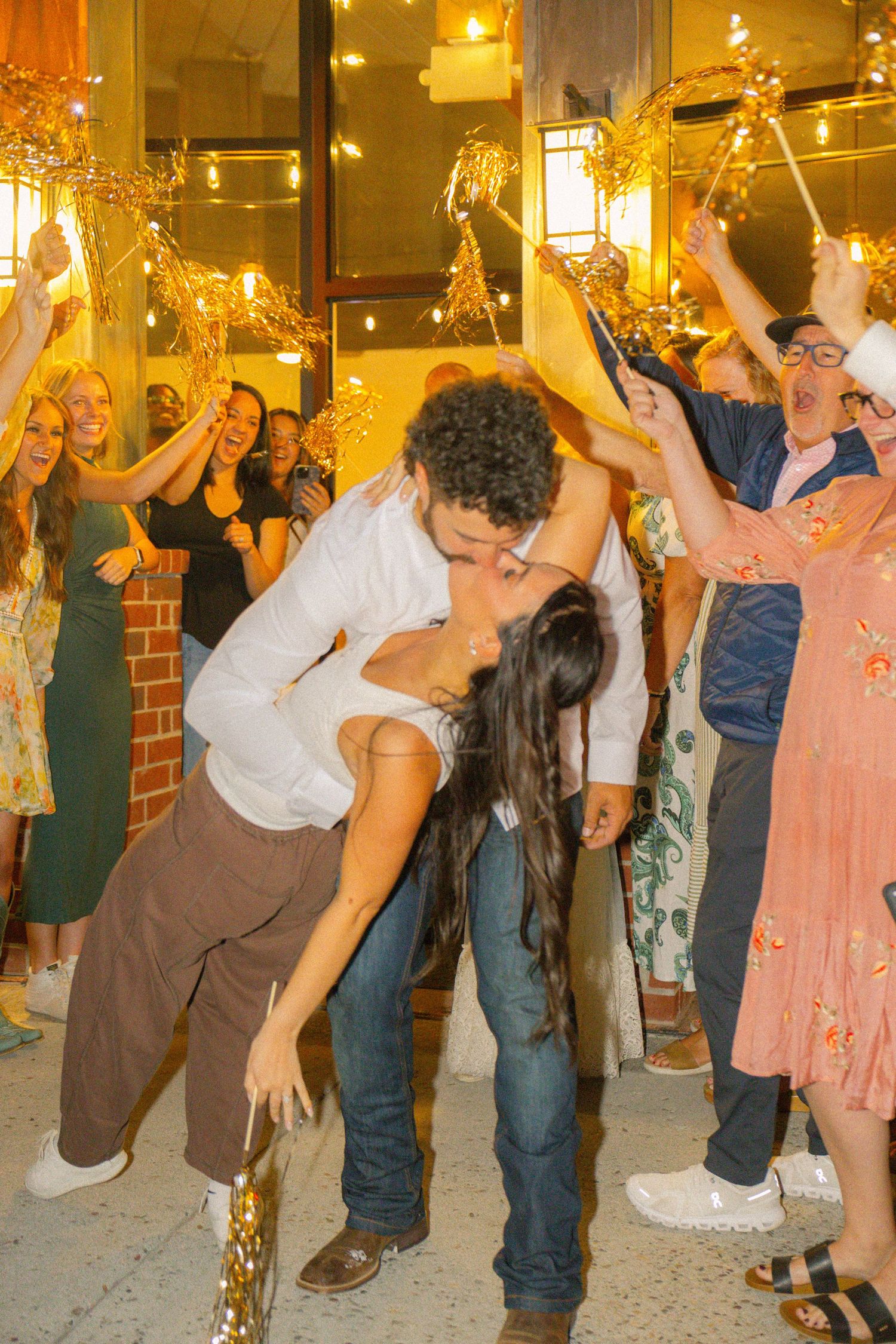 Couple shares romantic dip kiss surrounded by cheering guests holding sparklers at a festive celebration.