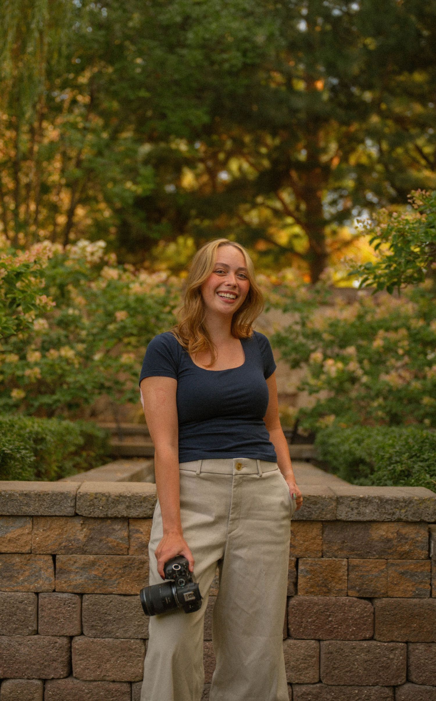 Smiling blonde woman holding a DSLR camera, wearing a navy top and khaki skirt, standing in a lush garden setting.