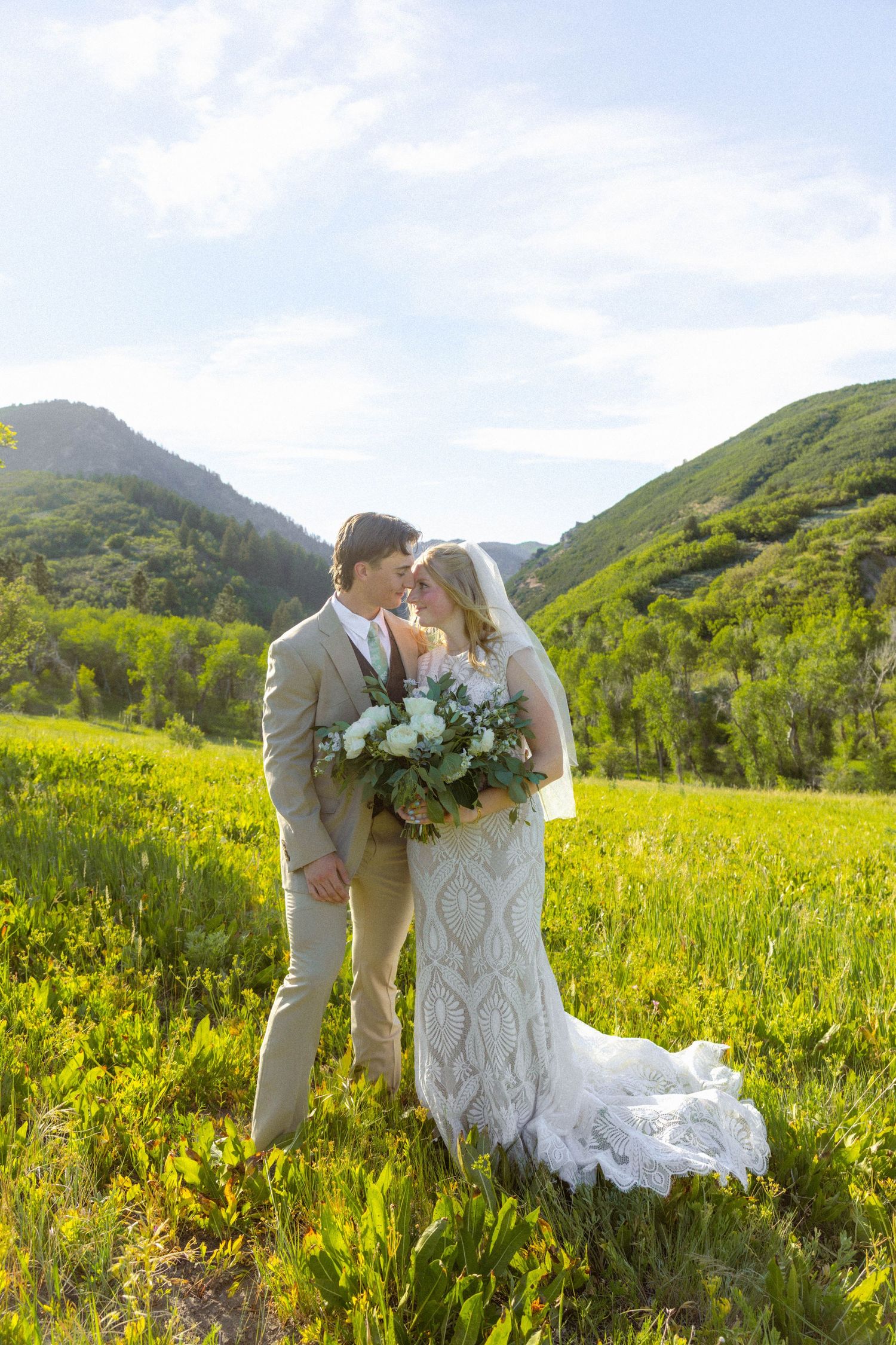 Bride and groom embrace in a lush green mountain meadow, bride holding white bouquet in elegant lace gown.