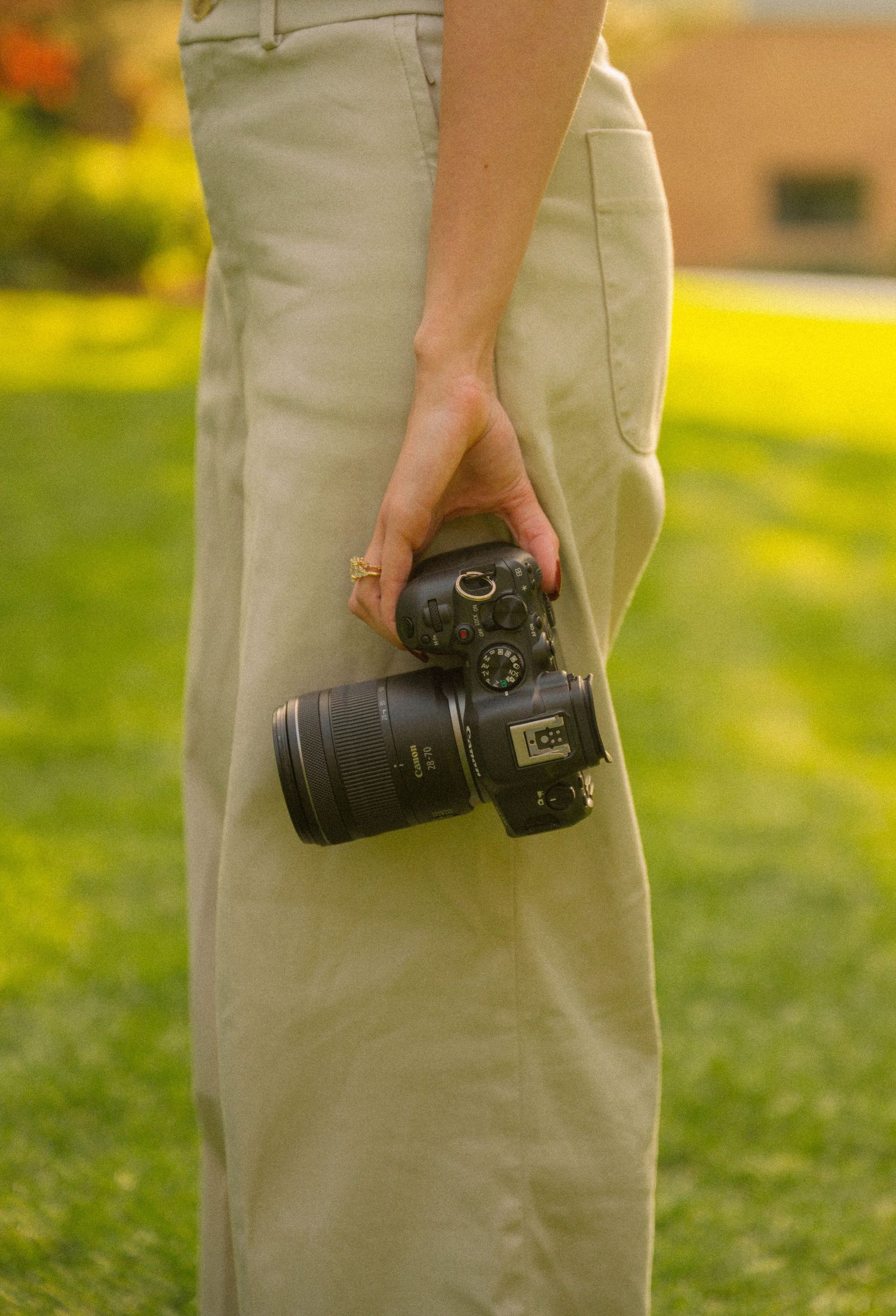 Person holding a Canon DSLR camera with zoom lens outdoors on green grass, wearing khaki pants and a gold ring.