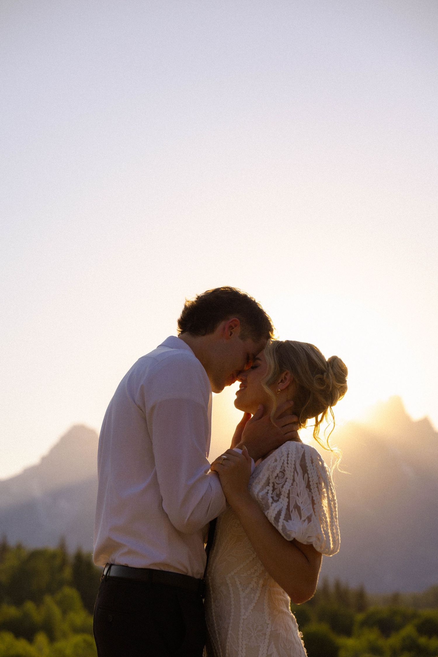 Couple sharing a romantic moment at sunset with mountain backdrop, bride in lace dress and groom in white shirt.