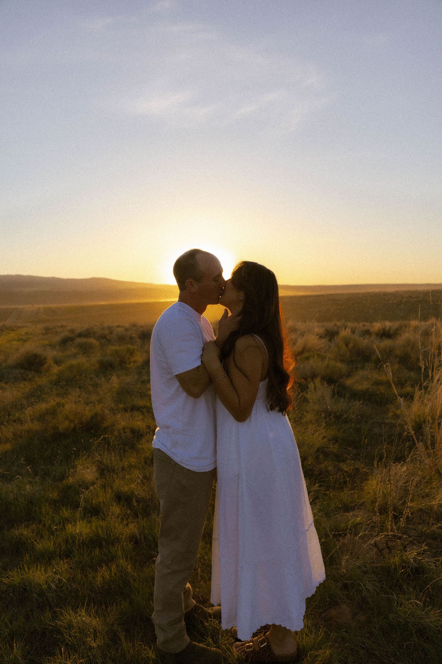 Couple sharing a romantic kiss at golden sunset in a grassy field, woman in white dress, warm backlit glow.