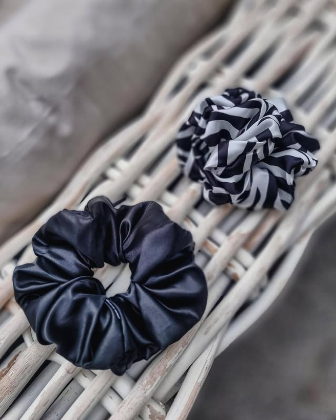 Two hair scrunchies on a wicker surface, one solid black satin and one black and white striped.