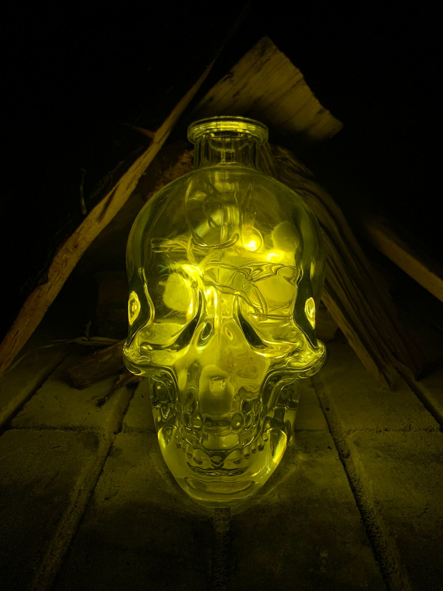 A crystal clear glass skull bottle glowing with yellow light on wooden surface in dark setting.