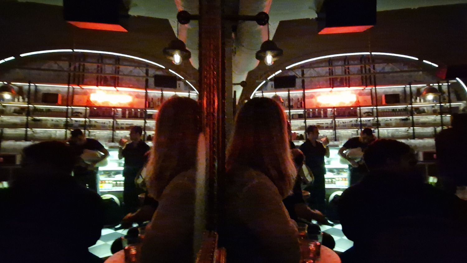A dark bar interior reflected in a mirror showing patrons sitting under warm red pendant lights.