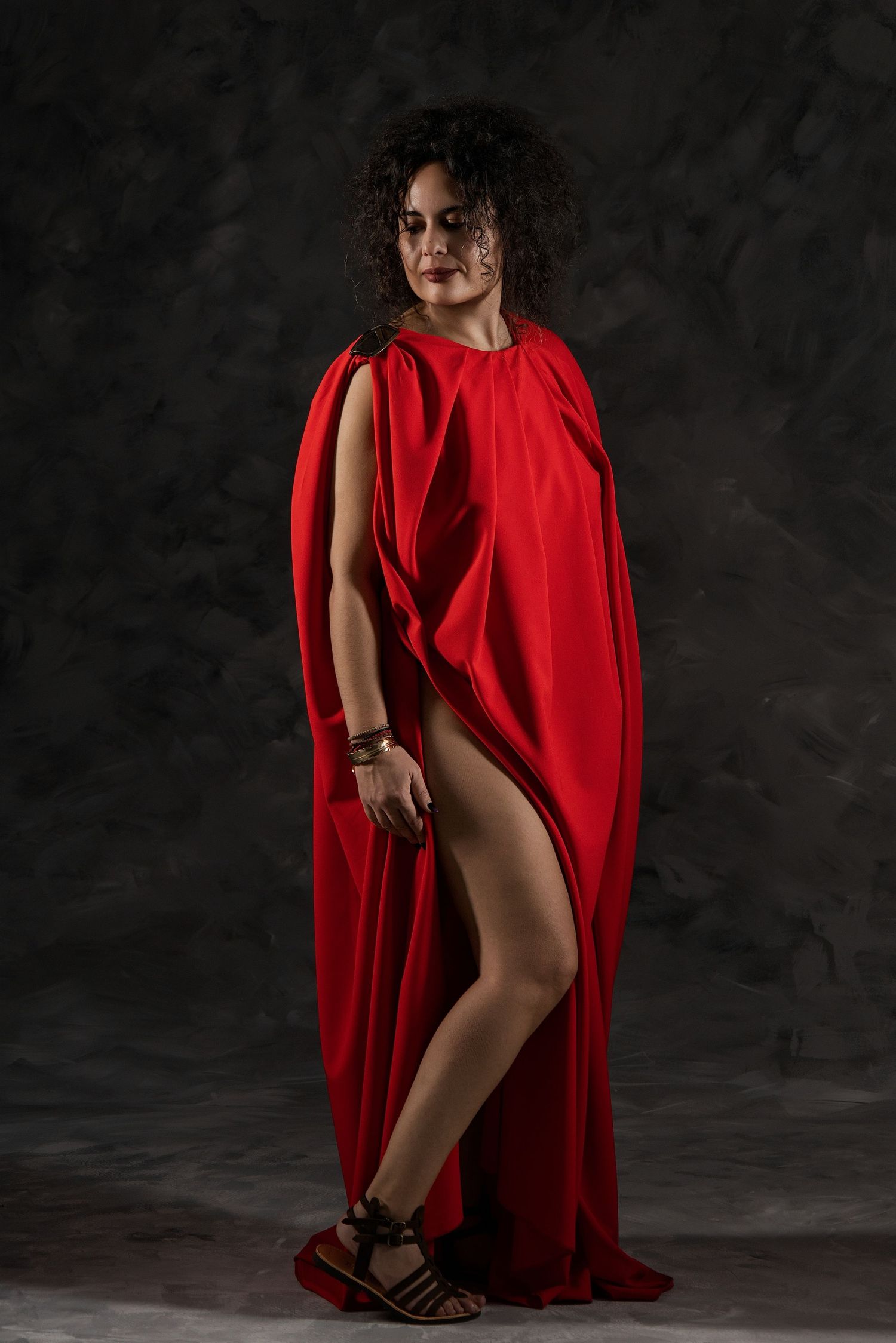 Woman in a dramatic red draped cape dress poses confidently against a dark textured background.