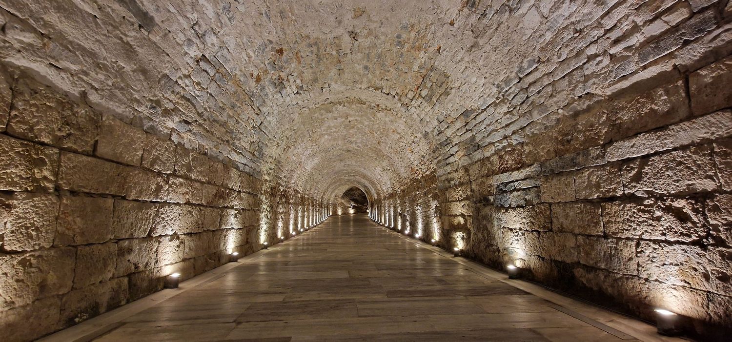 Ancient stone tunnel corridor with arched ceiling illuminated by warm floor lights leading to a vanishing point.