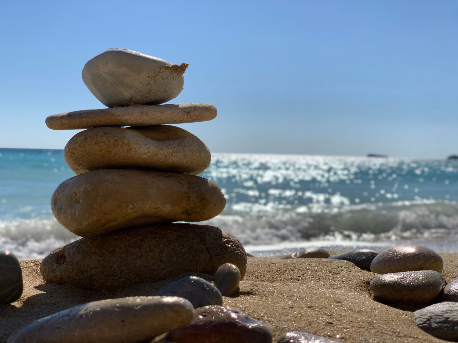 Balanced rock cairn stack on sandy beach shore with blue ocean waves and clear sky behind.