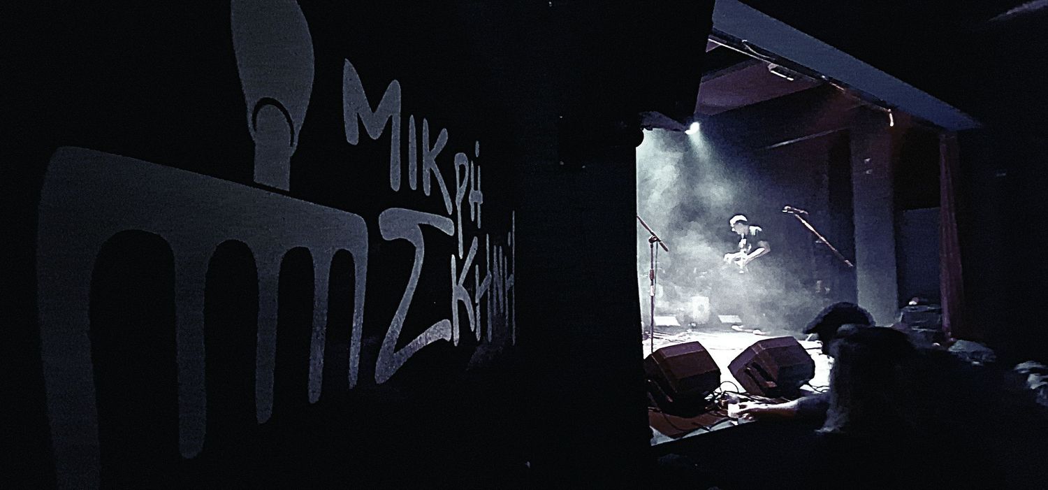 Dark concert venue interior with stage lighting and Mikri Zikara venue sign on the wall.