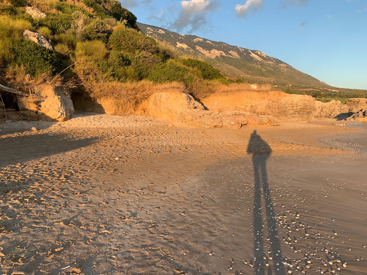 Long shadow cast on sandy beach at golden hour with eroded cliffs and mountain backdrop.