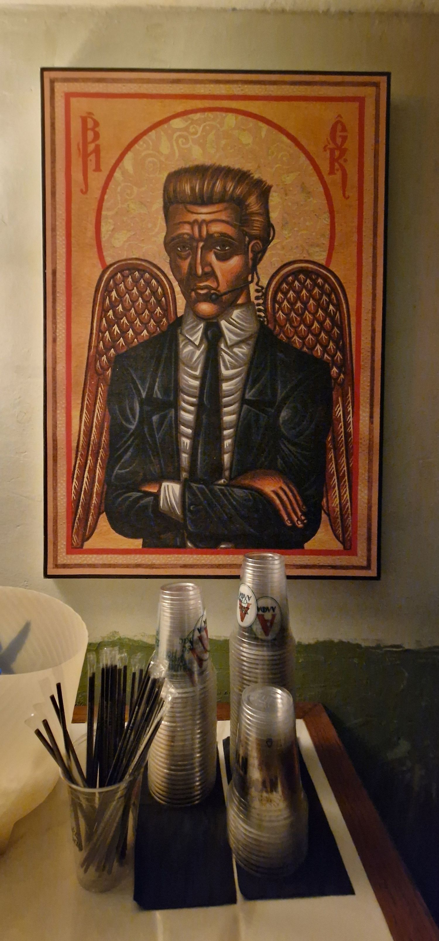 Vintage icon-style painting of a suited angel with crossed arms hanging above stacked plastic cups and stirrers.