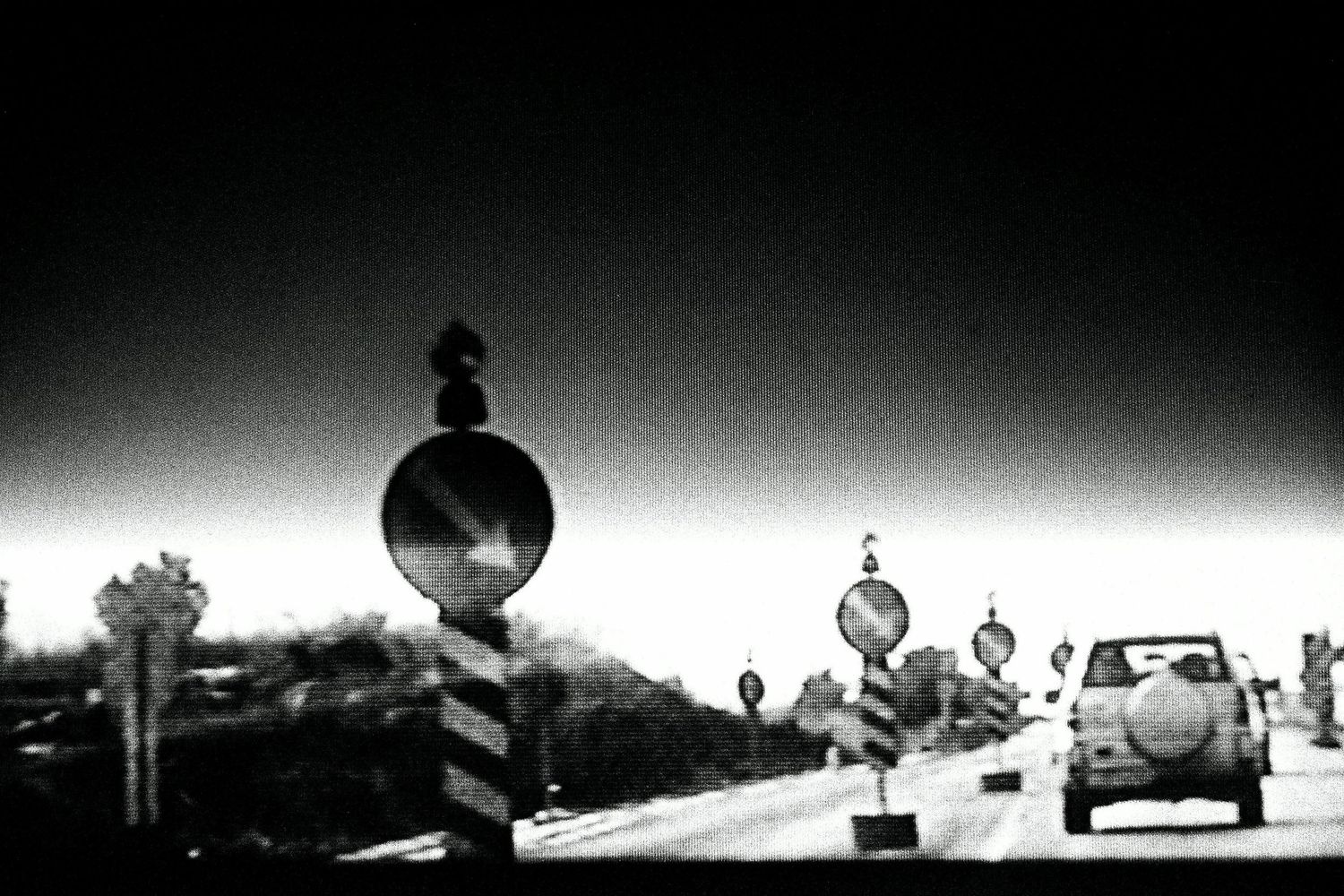 Black and white photo of road construction signs and a car on a highway under a bright sky.
