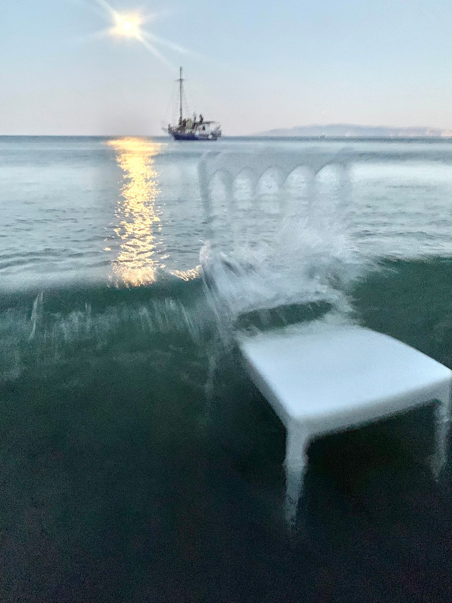 White plastic chair submerged in ocean waves at sunset with a sailing boat visible on the horizon.
