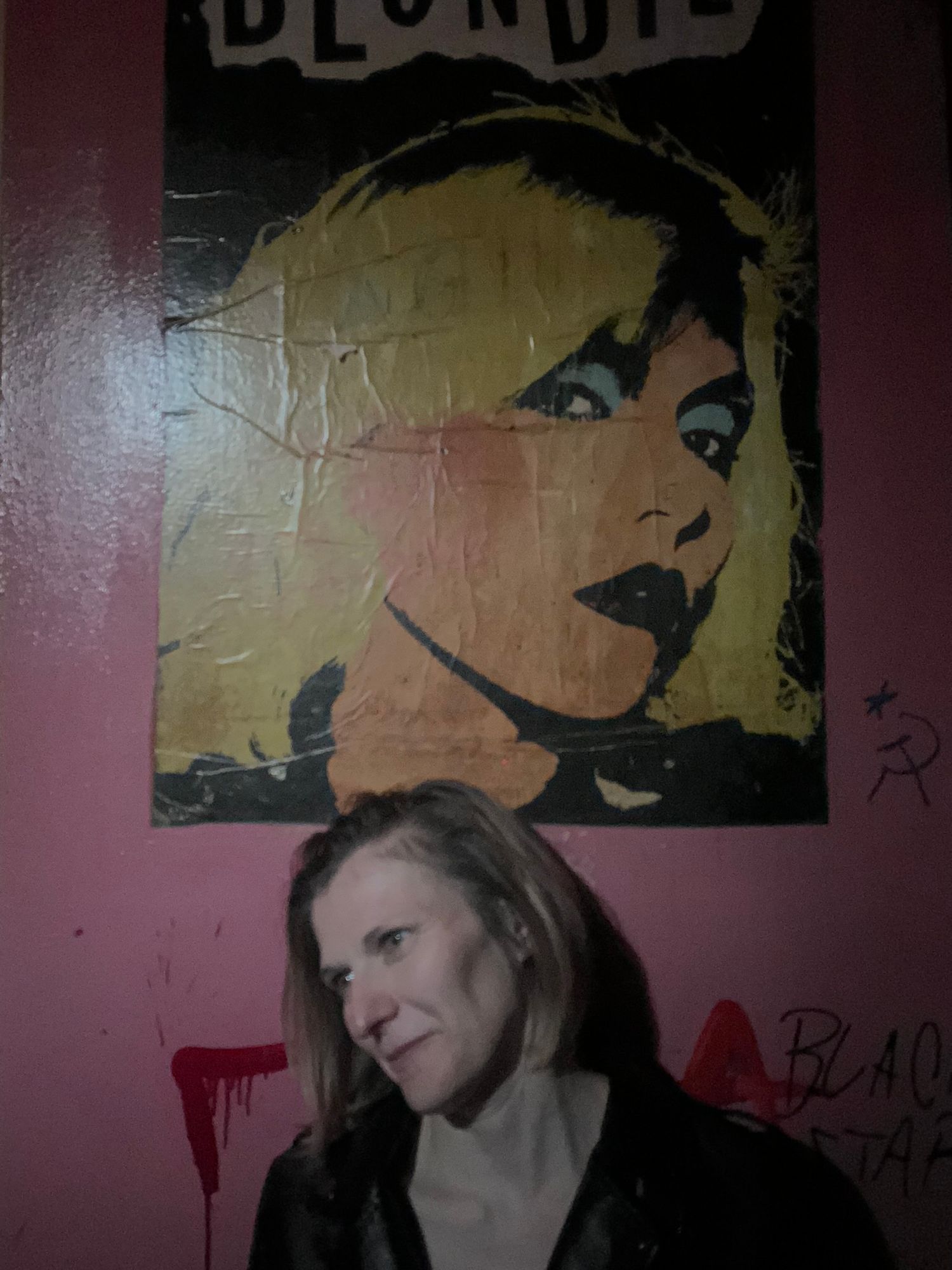 Woman standing beneath a vintage Blondie band poster on a pink graffiti-covered wall in a bar.