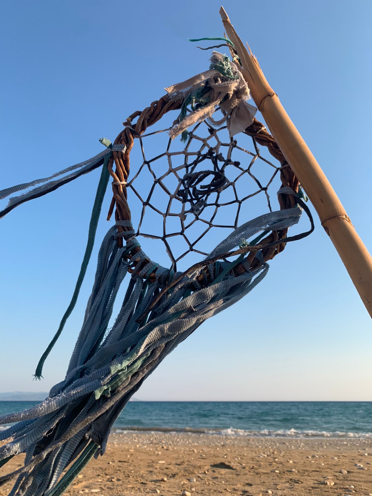 A rustic dreamcatcher with woven web hangs on a bamboo pole at a sandy beach under a clear blue sky.