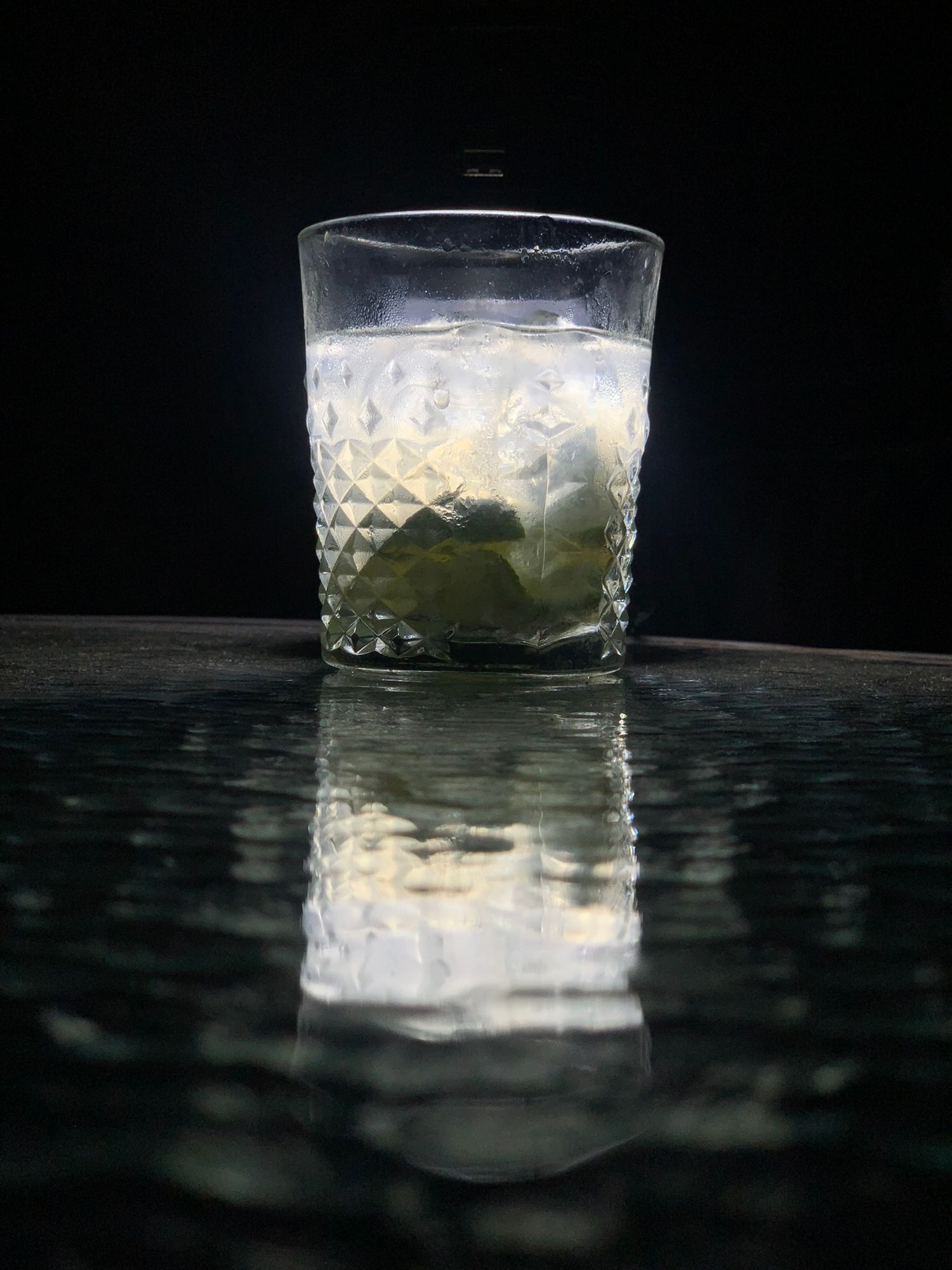 Crystal cut glass with lime and ice cocktail reflecting on a dark wet surface against black background.