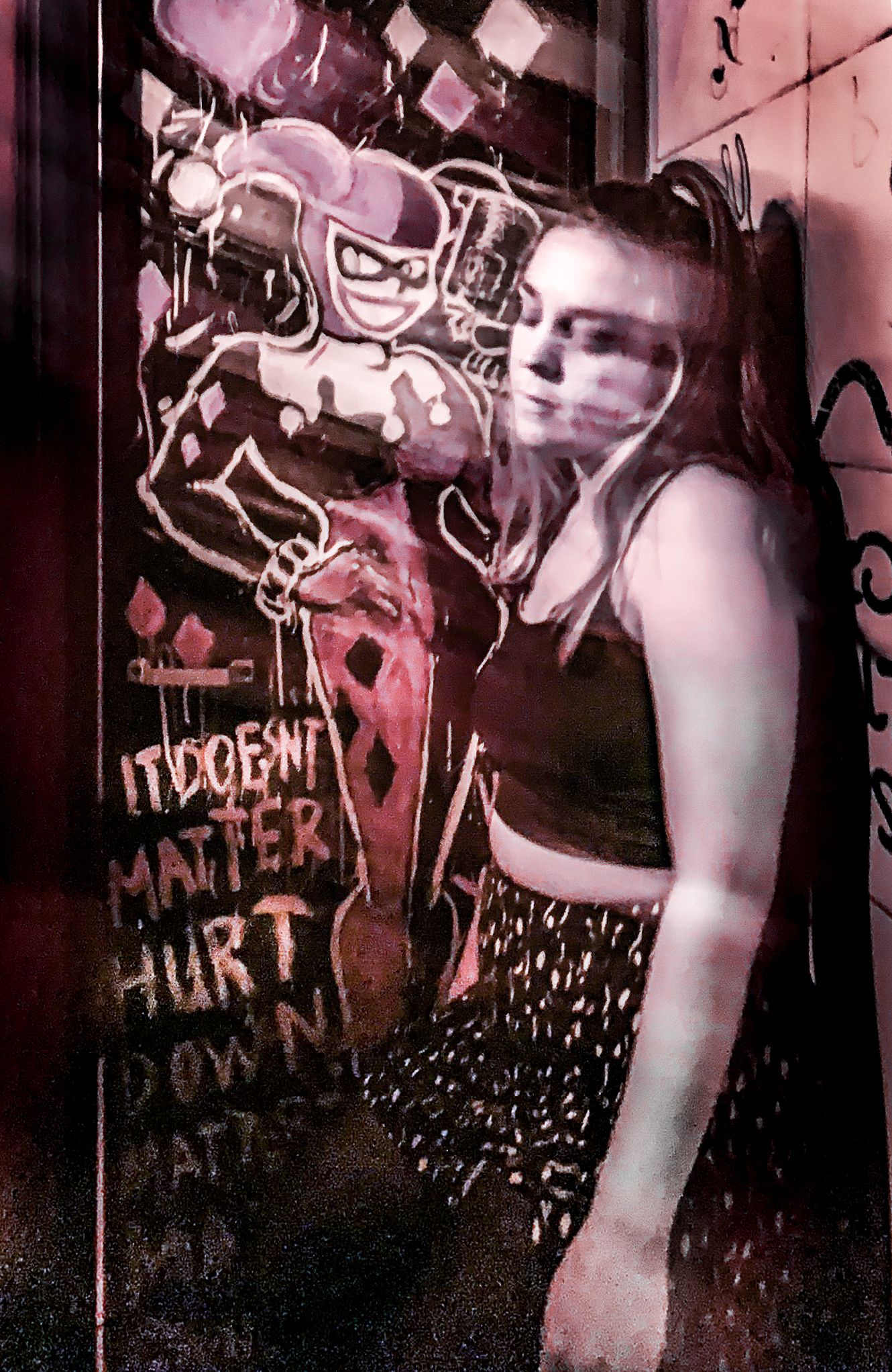 Young woman posing beside colorful Harley Quinn graffiti mural on an urban wall.