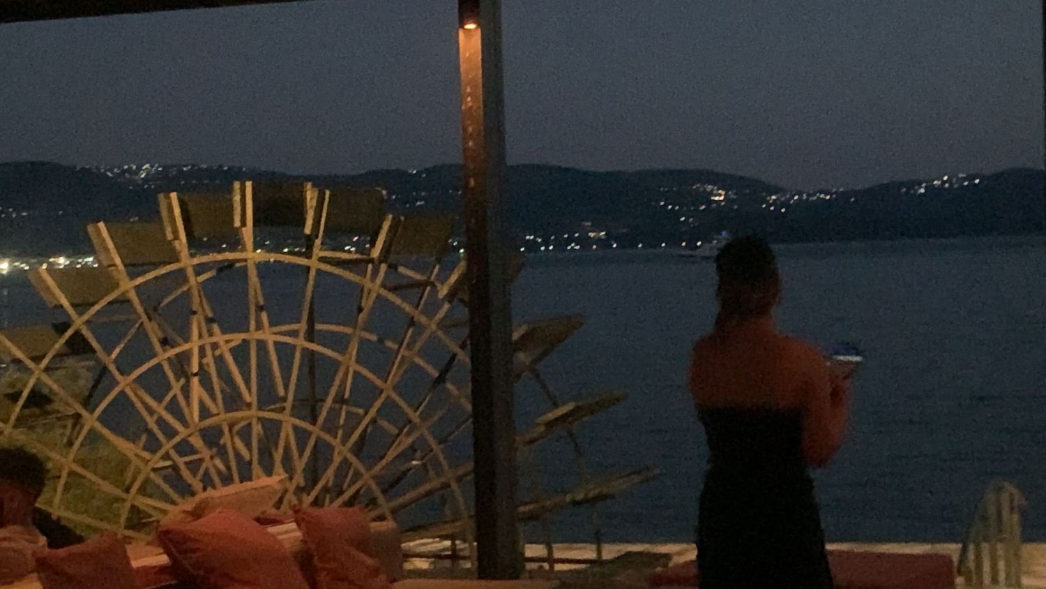 Person standing on a terrace overlooking a calm sea at dusk next to a large wicker decoration.