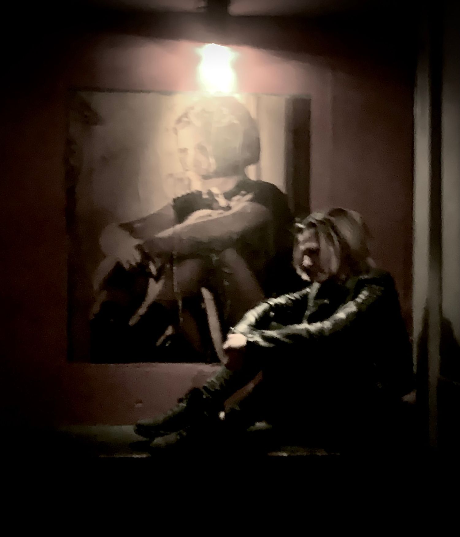 Person sitting on floor beneath a dramatic black and white portrait photograph in a dimly lit room.