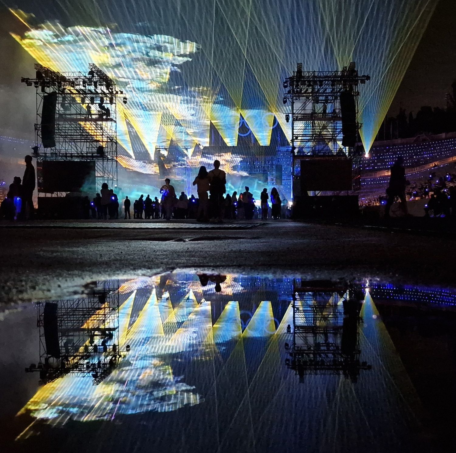 Spectacular laser light show reflects in a puddle at an outdoor music festival stage at night.
