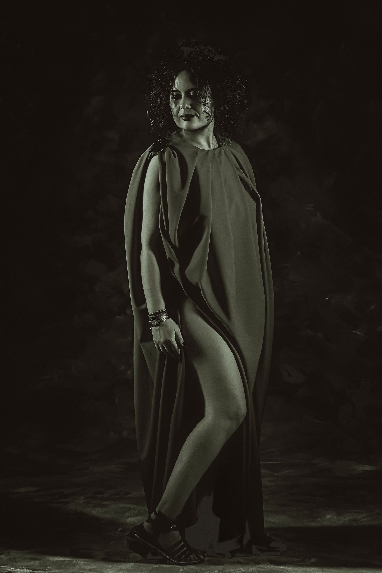 Moody black and white portrait of a curly-haired woman in a flowing draped garment and sandals.