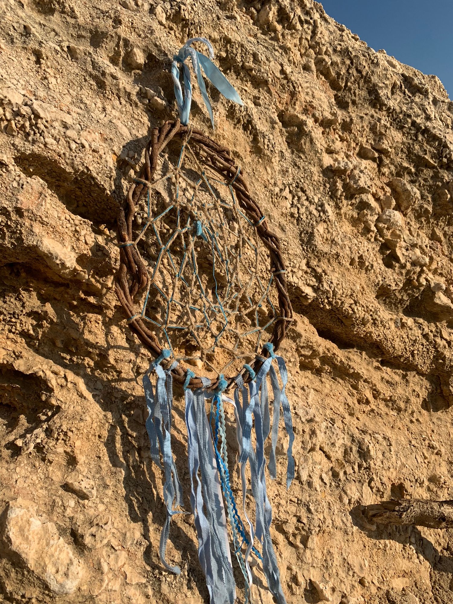 Handmade dream catcher with blue ribbons and beads hanging on a sandy rock cliff.