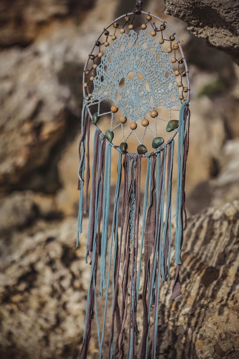 A boho blue lace dreamcatcher with wooden beads and long colorful fringe hangs against a rocky surface.