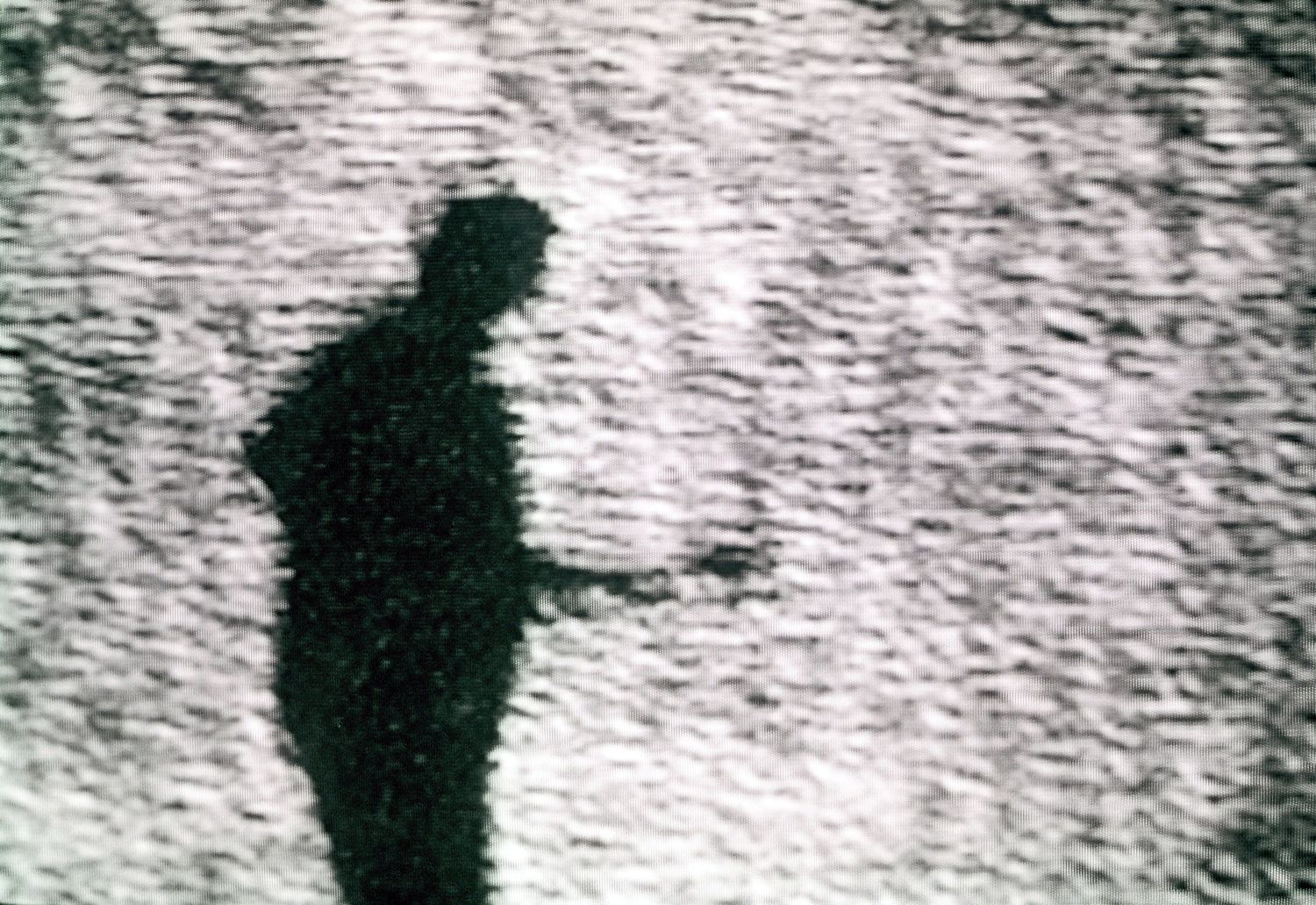 Grainy black and white shadow of a lone person standing against a textured light background.