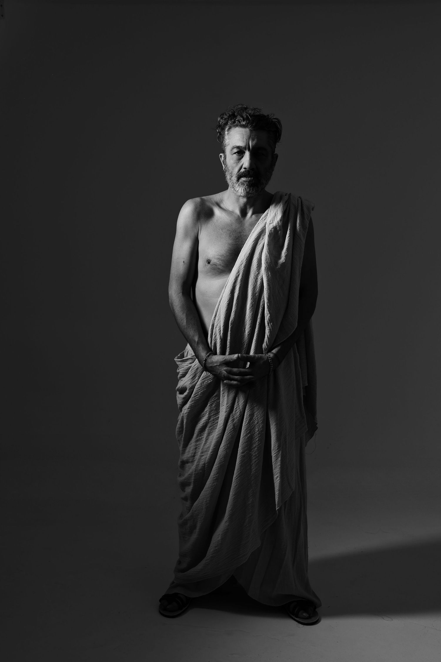 Black and white portrait of a middle-aged man draped in a toga, standing against a dark studio background.