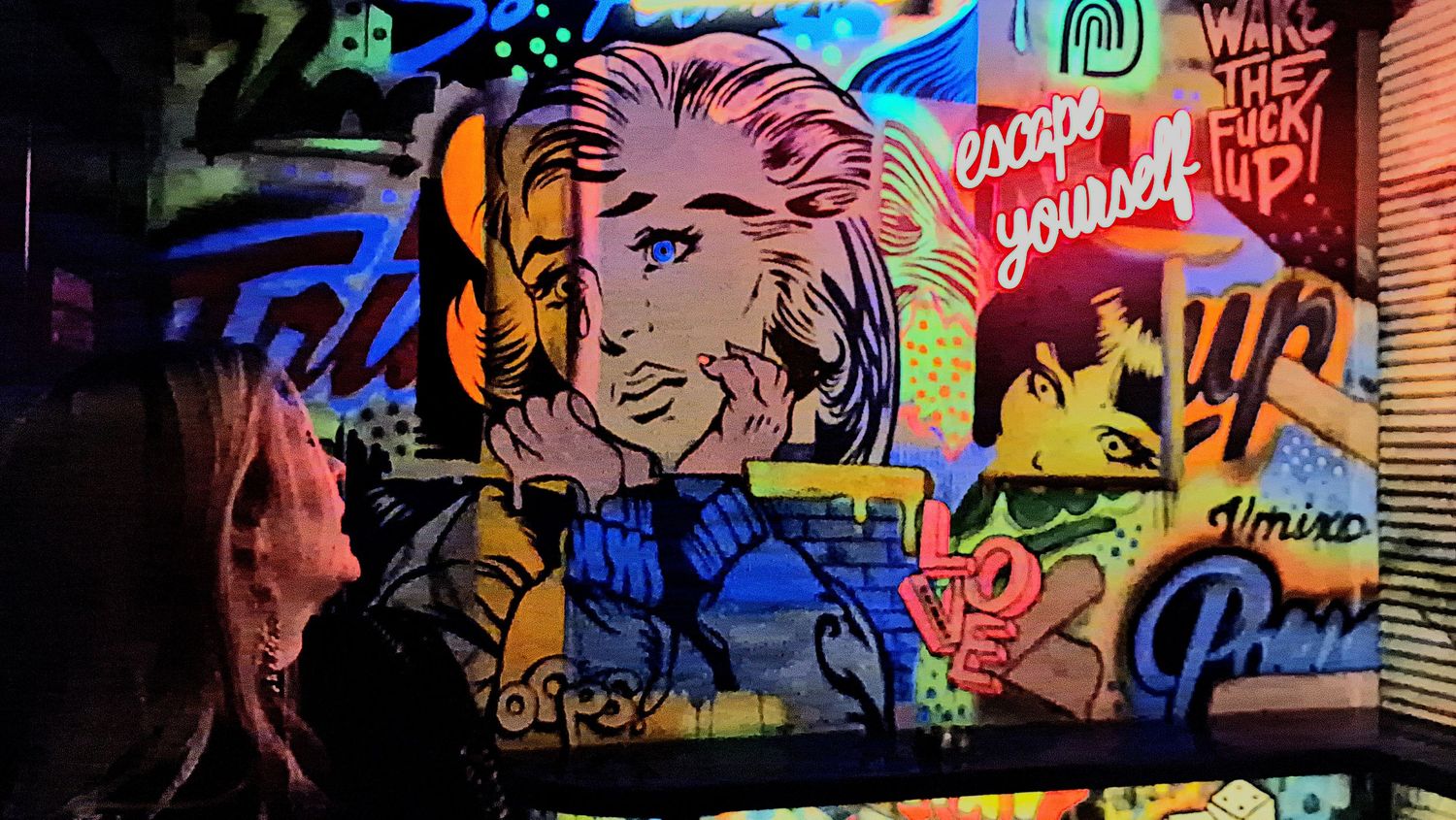 Woman viewing vibrant pop art graffiti mural with comic book style figures and colorful text on wall.