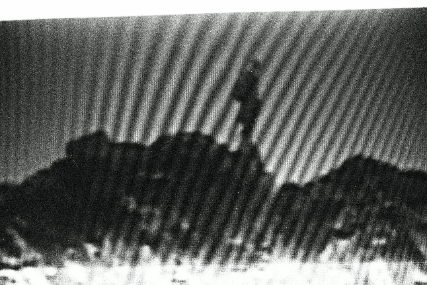 A grainy black and white photo of a lone silhouetted figure standing on a rocky hilltop at dusk.