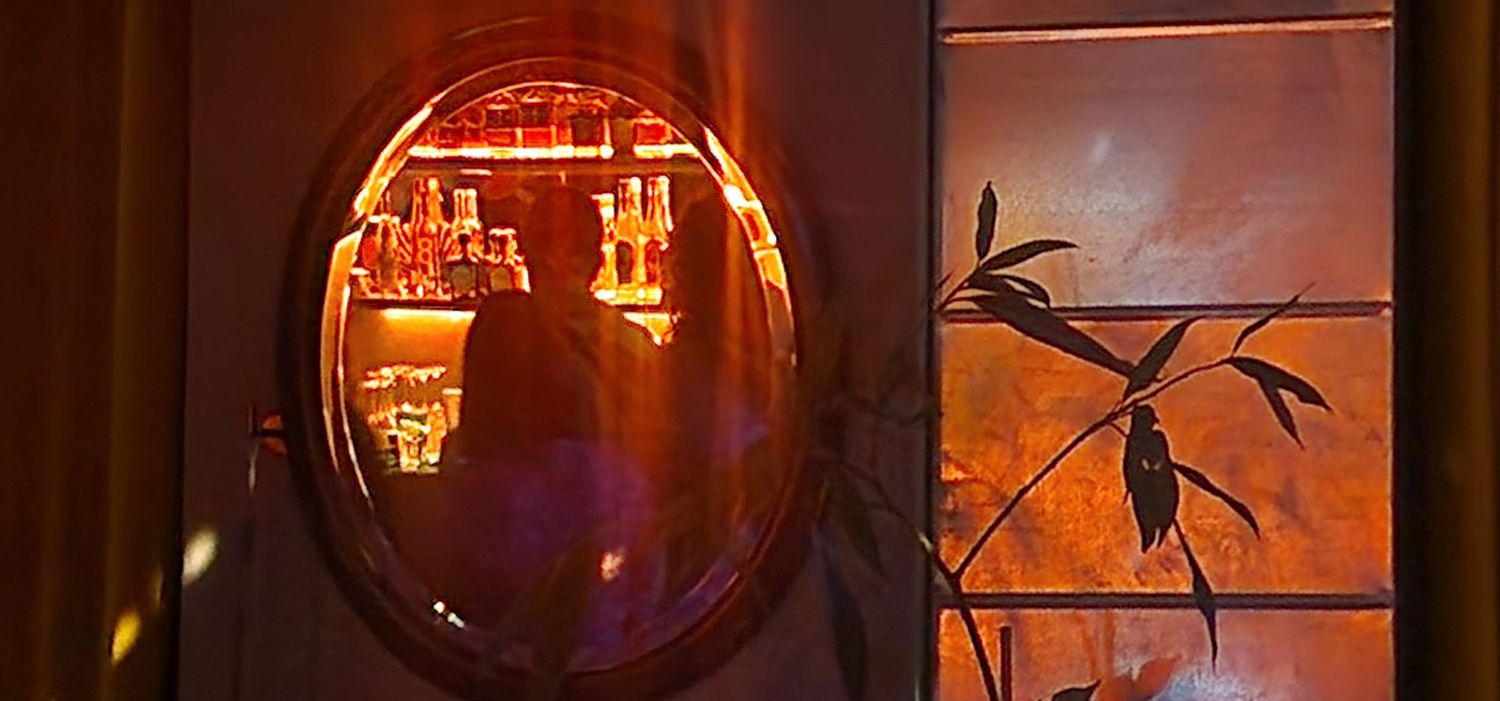 Warm orange light reflects through an oval mirror revealing a bar interior beside a window with plant silhouettes.