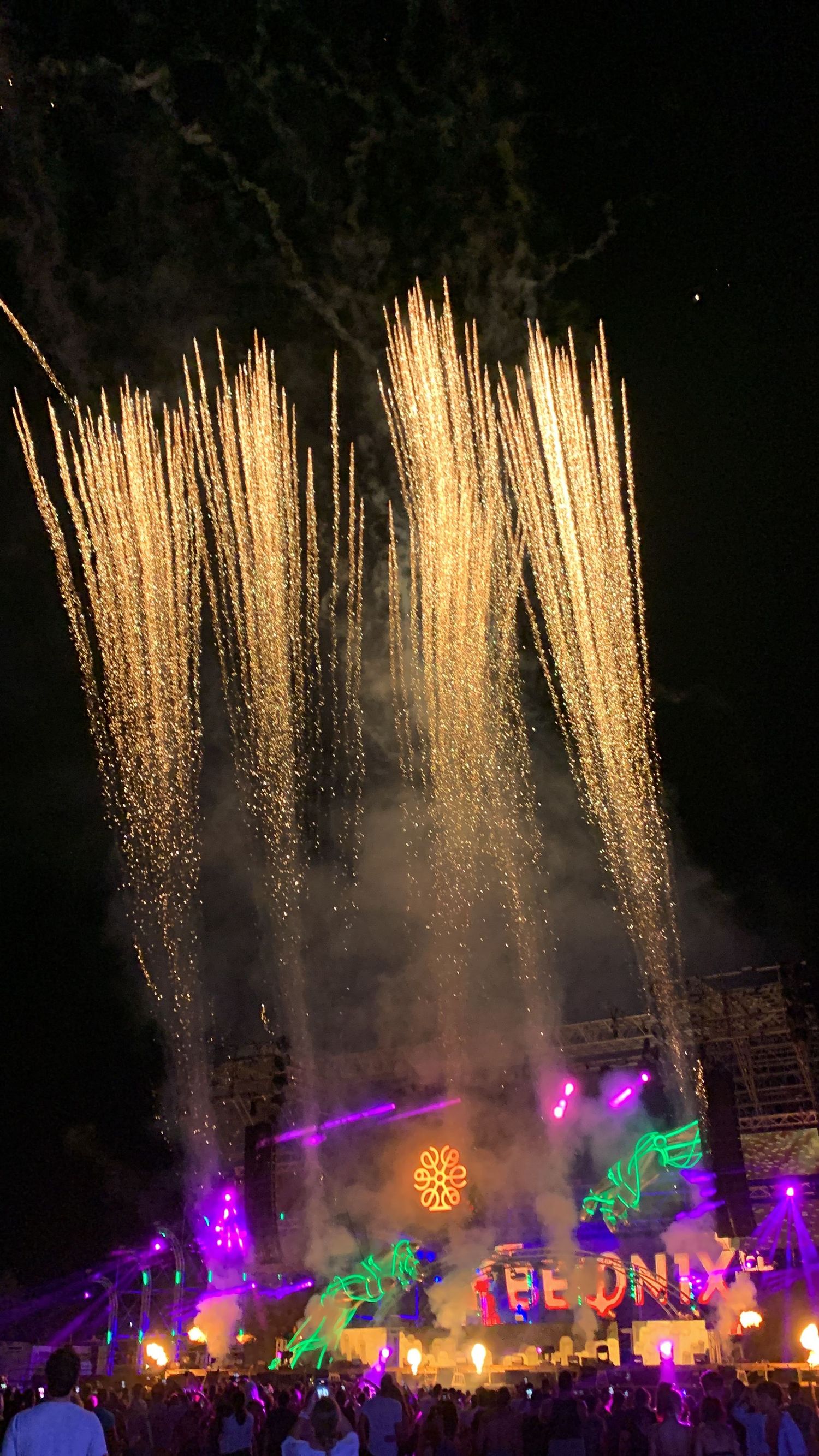 Golden fireworks shooting into the night sky above a colorful neon music festival stage.