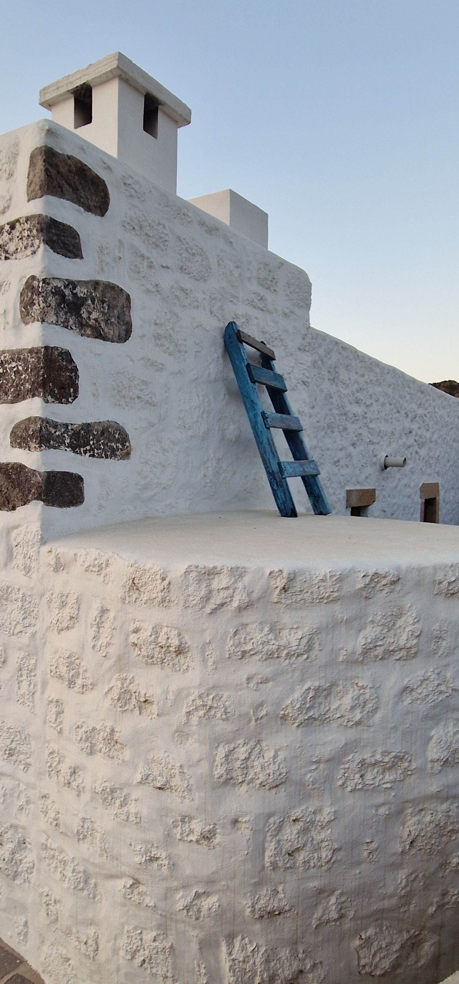 White-washed stone building with blue wooden ladder leaning against wall under clear sky.