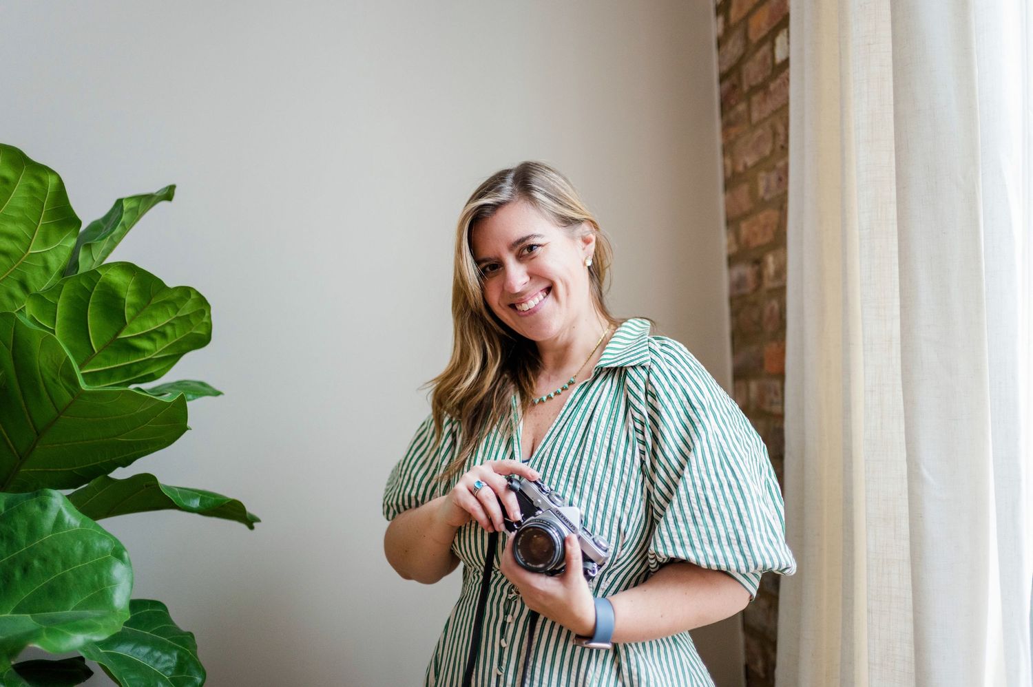 A photographer in a striped button-down shirt holds a camera while standing next to indoor plants.