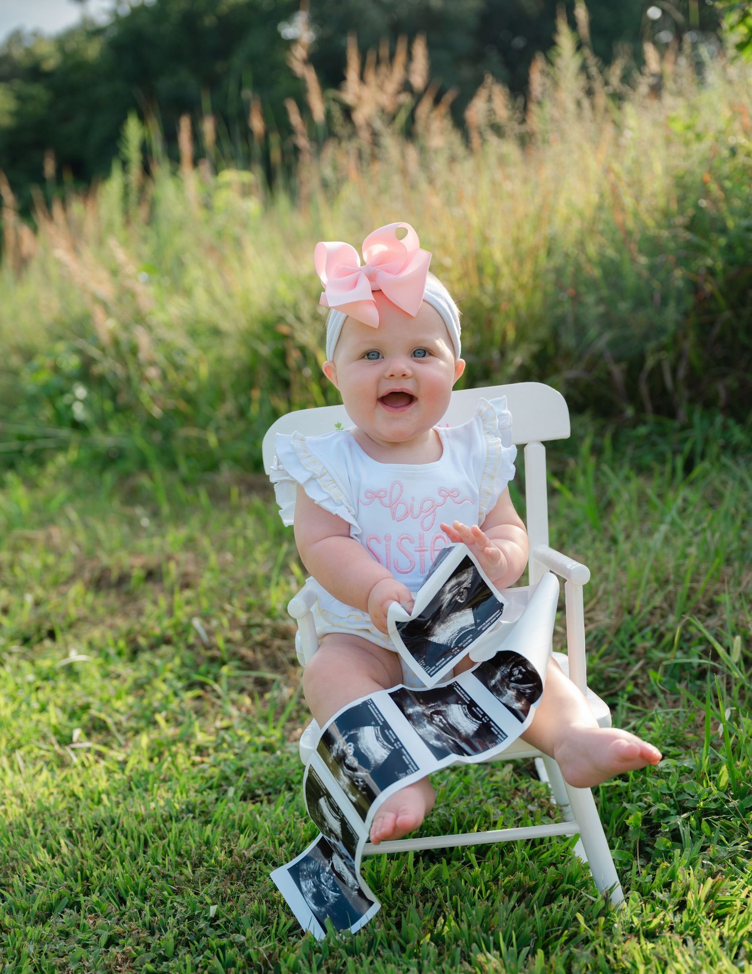 Baby in white dress sits on chair outdoors holding photo strips with pink bow accessory.